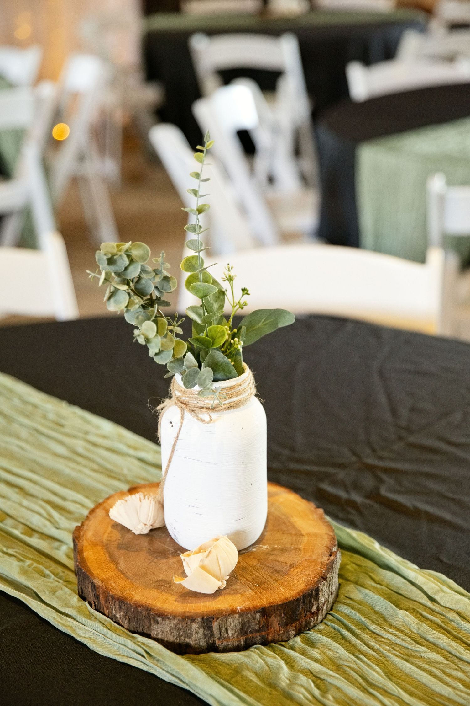 Rustic wedding centerpiece with white mason jar holding eucalyptus on wood slice with green table runner.