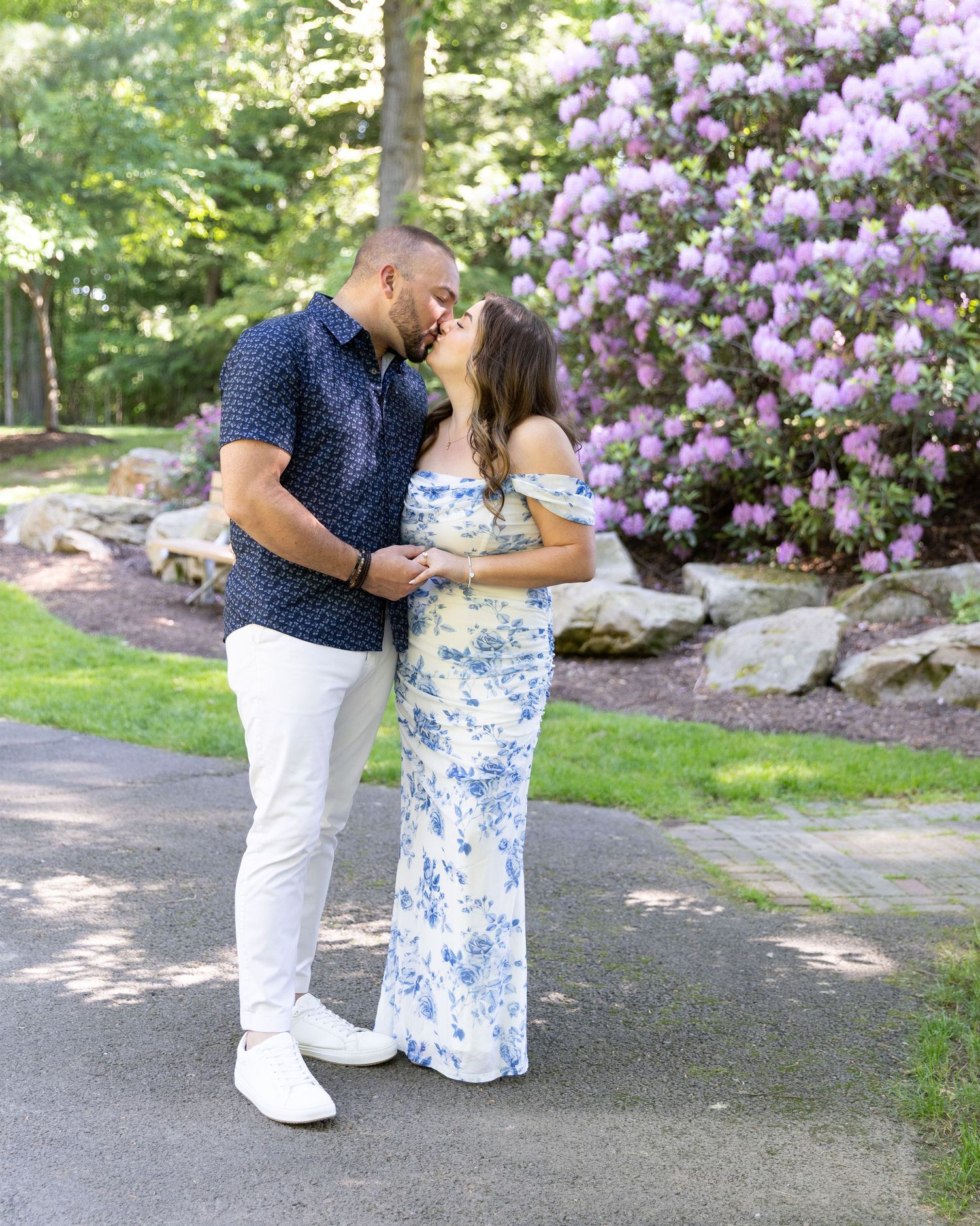 Couple sharing a romantic kiss in a garden path surrounded by blooming purple rhododendrons and lush green trees.