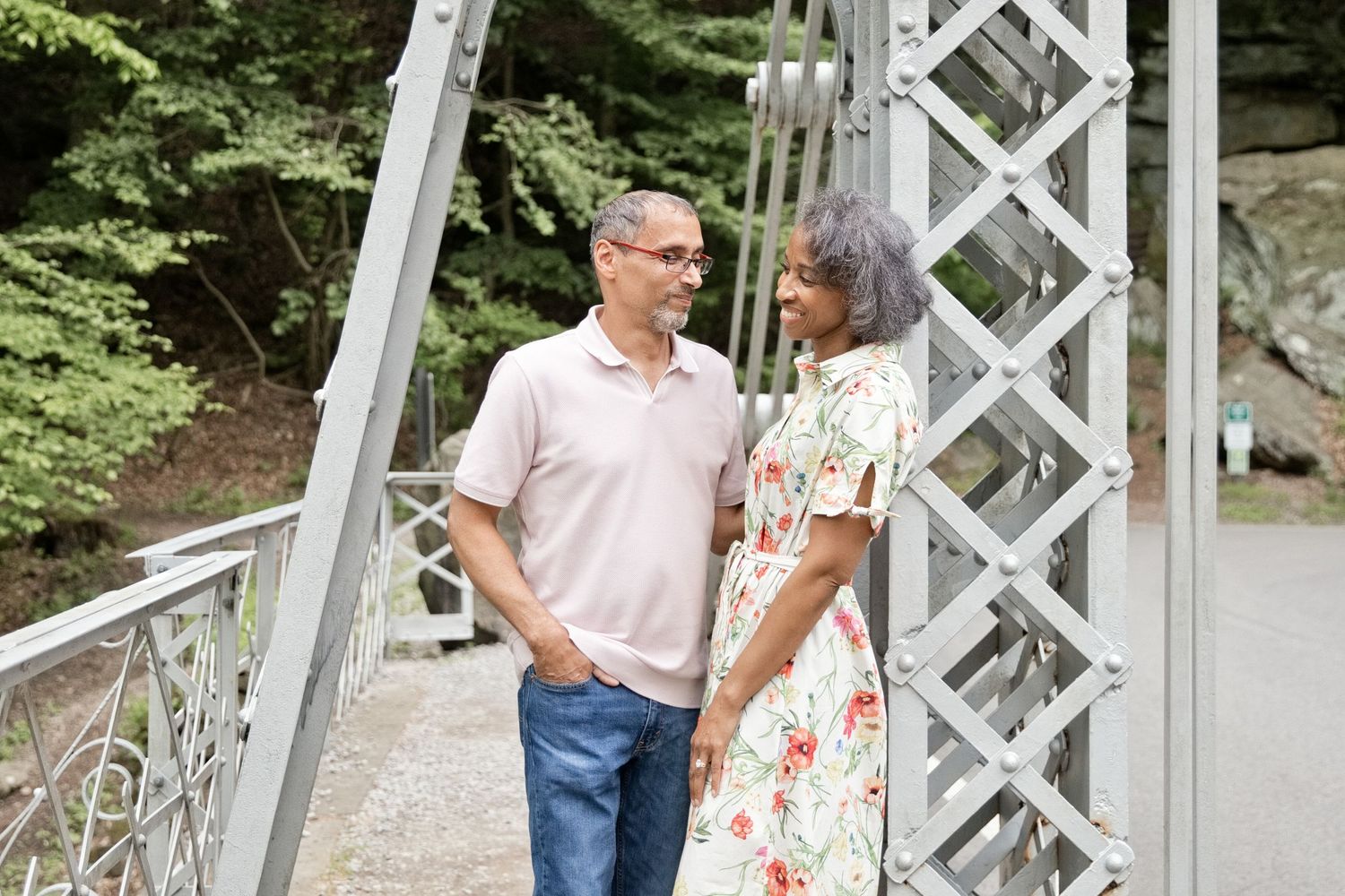 A middle-aged couple stands on a white metal bridge surrounded by lush green trees, sharing a loving gaze.
