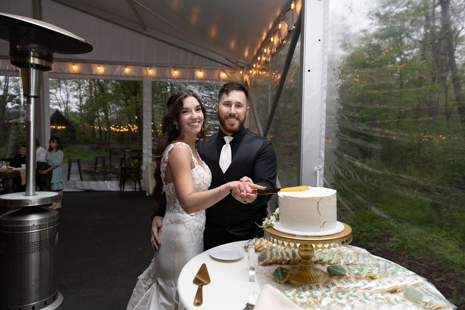 Bride and groom smiling while cutting their white wedding cake under a tented outdoor reception with string lights.