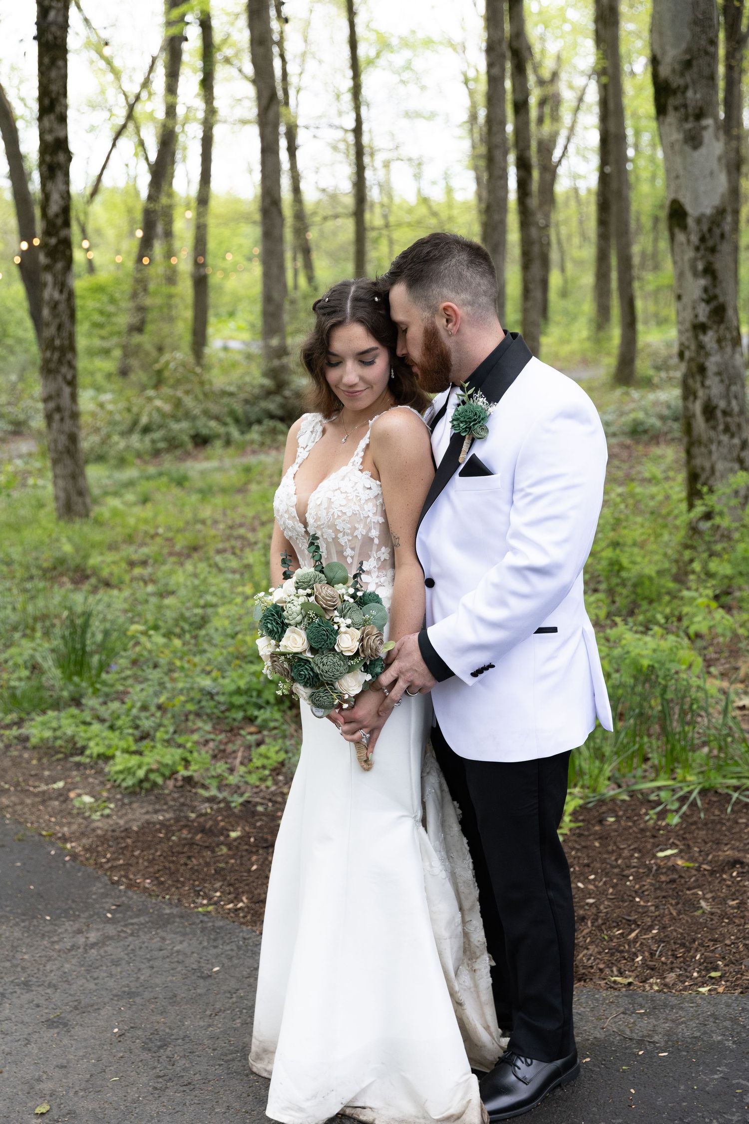 Bride and groom embracing in a lush green forest, bride holding a white and green bouquet in a white lace gown.