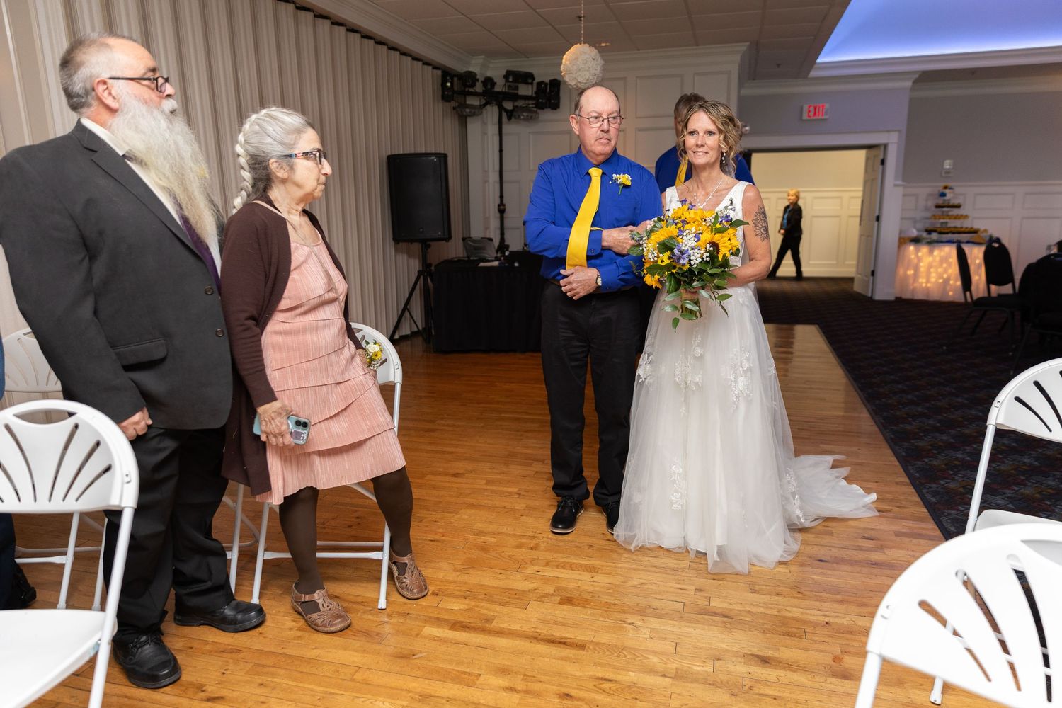 Bride in white gown holding yellow flowers walks with man in blue shirt past elderly couple at indoor wedding reception.