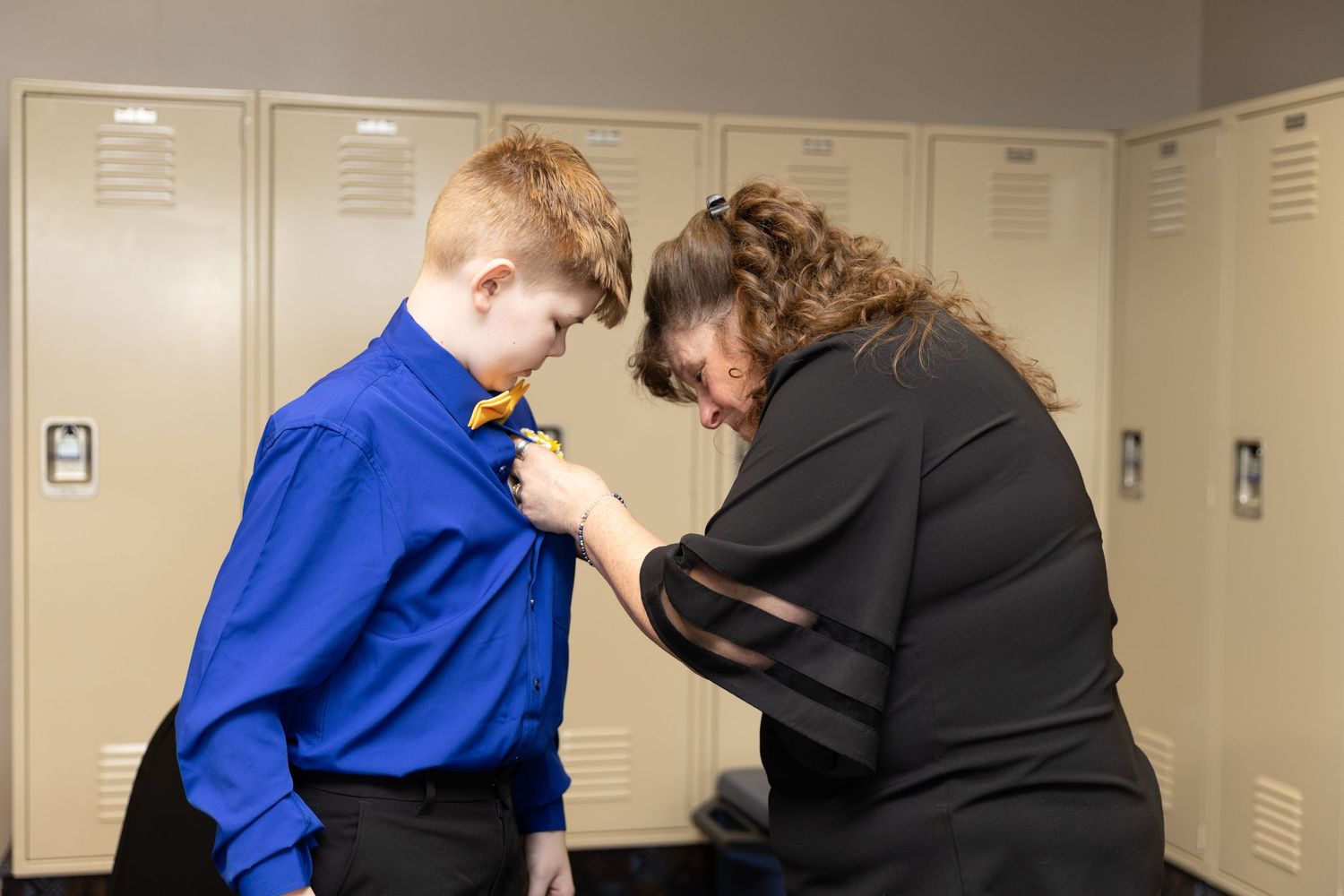Woman helping young boy in blue shirt adjust yellow bow tie near school lockers.