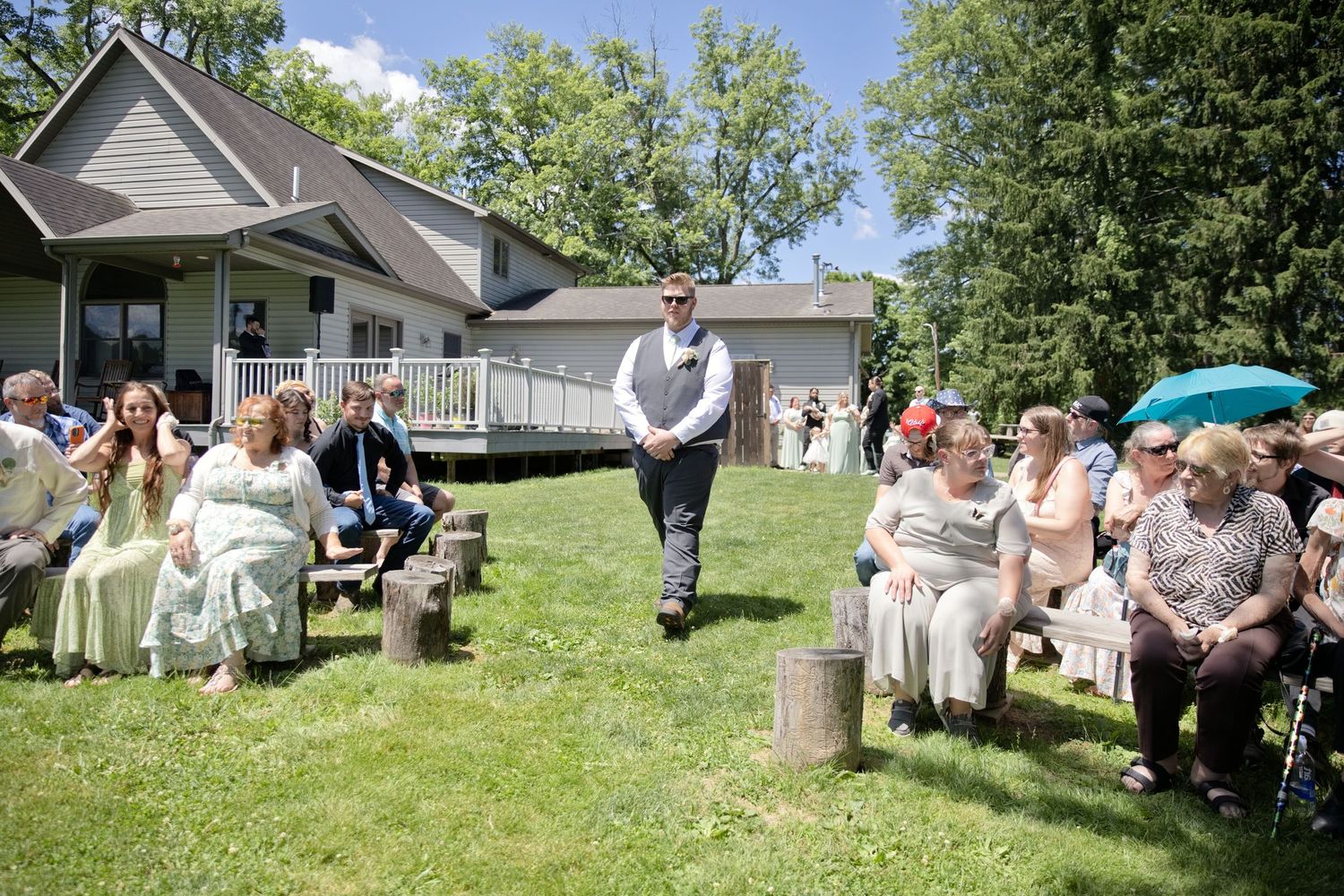 Outdoor backyard wedding ceremony with guests seated on logs, groom walking down the aisle toward a house on a sunny day.