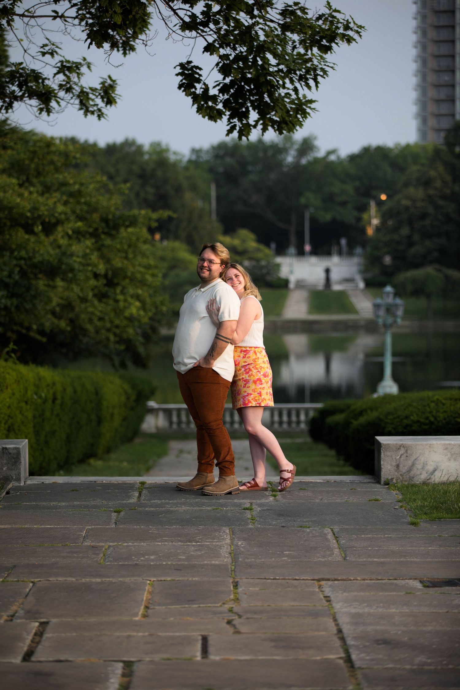 A romantic couple embraces on a stone pathway in a formal garden with trees and fountains in the background.