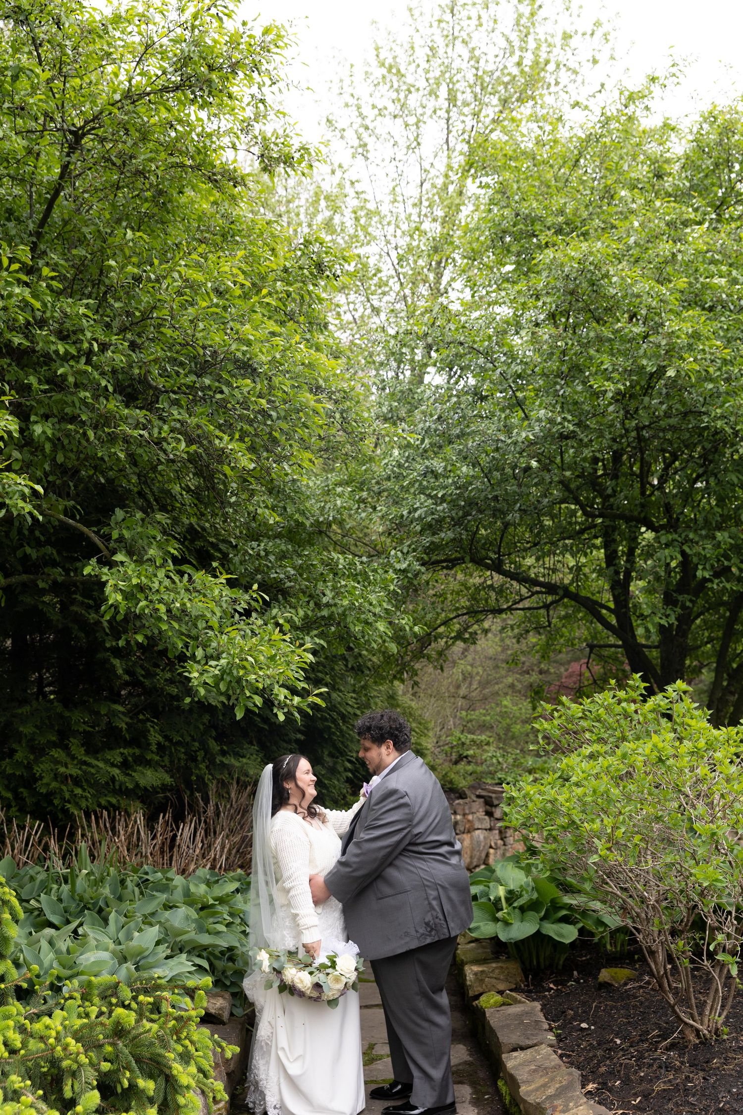 Bride and groom share a romantic moment in a lush green garden, bride holding a white bouquet in a flowing gown.