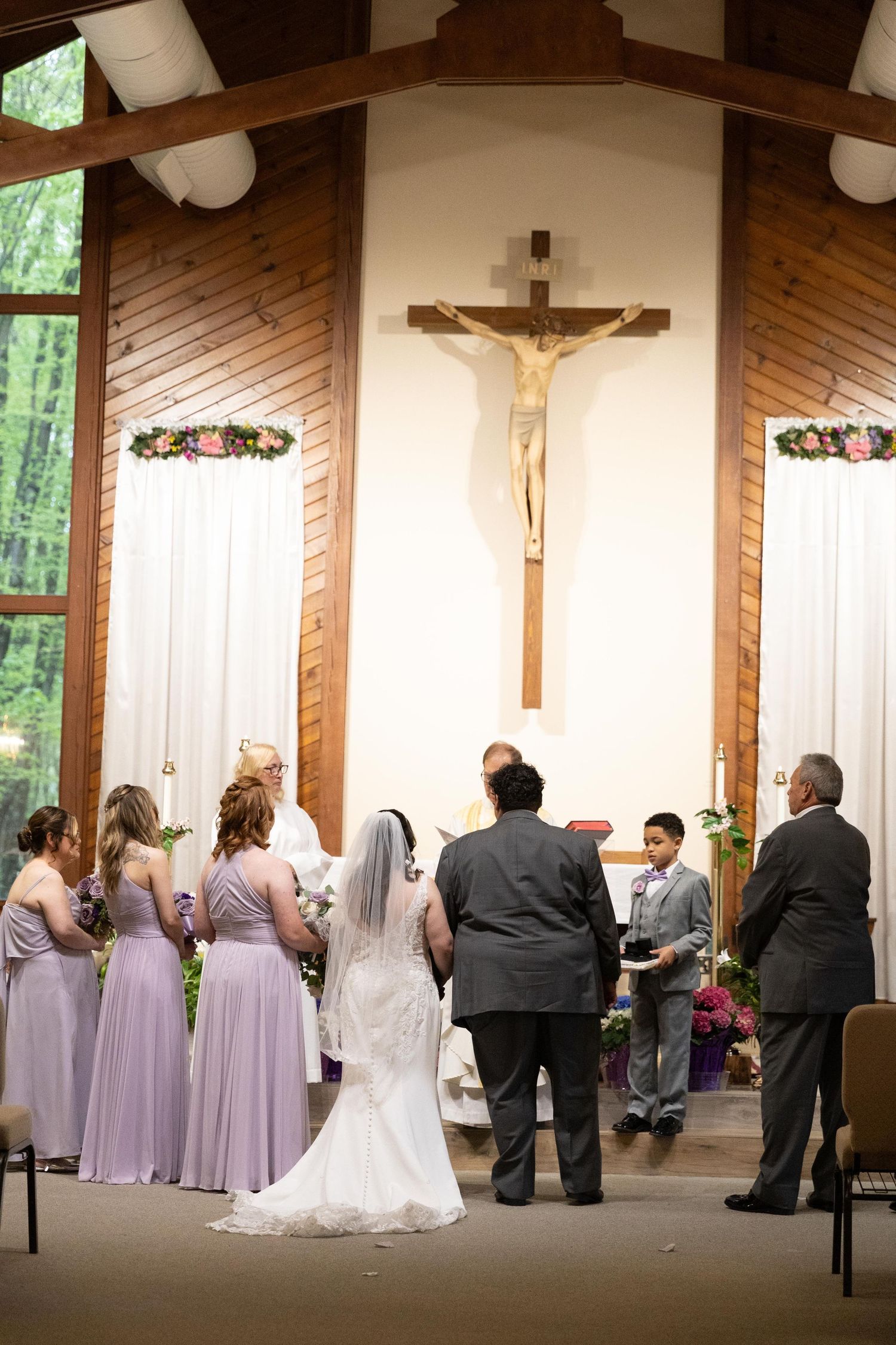 Catholic wedding ceremony at altar with bride, groom, bridesmaids in lavender dresses, and crucifix backdrop in wooden chapel.