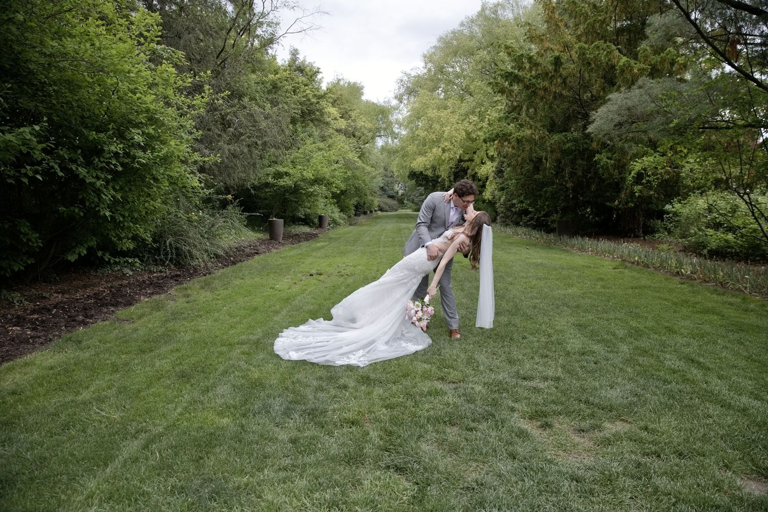 Groom dips bride in white gown and veil on lush green lawn surrounded by trees during outdoor wedding ceremony.
