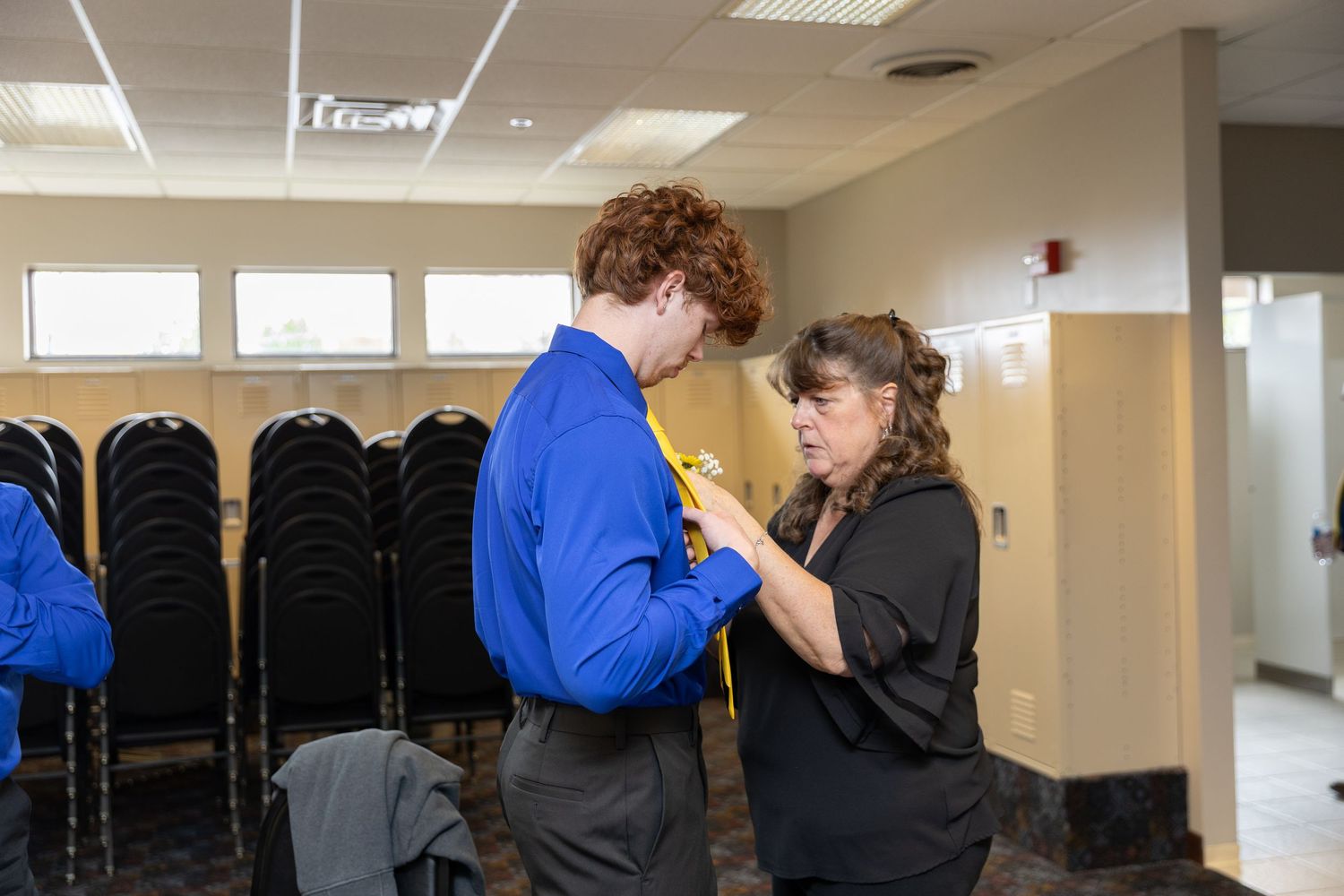A woman helps a young man with a yellow tie in a blue shirt inside a formal event venue with black chairs.