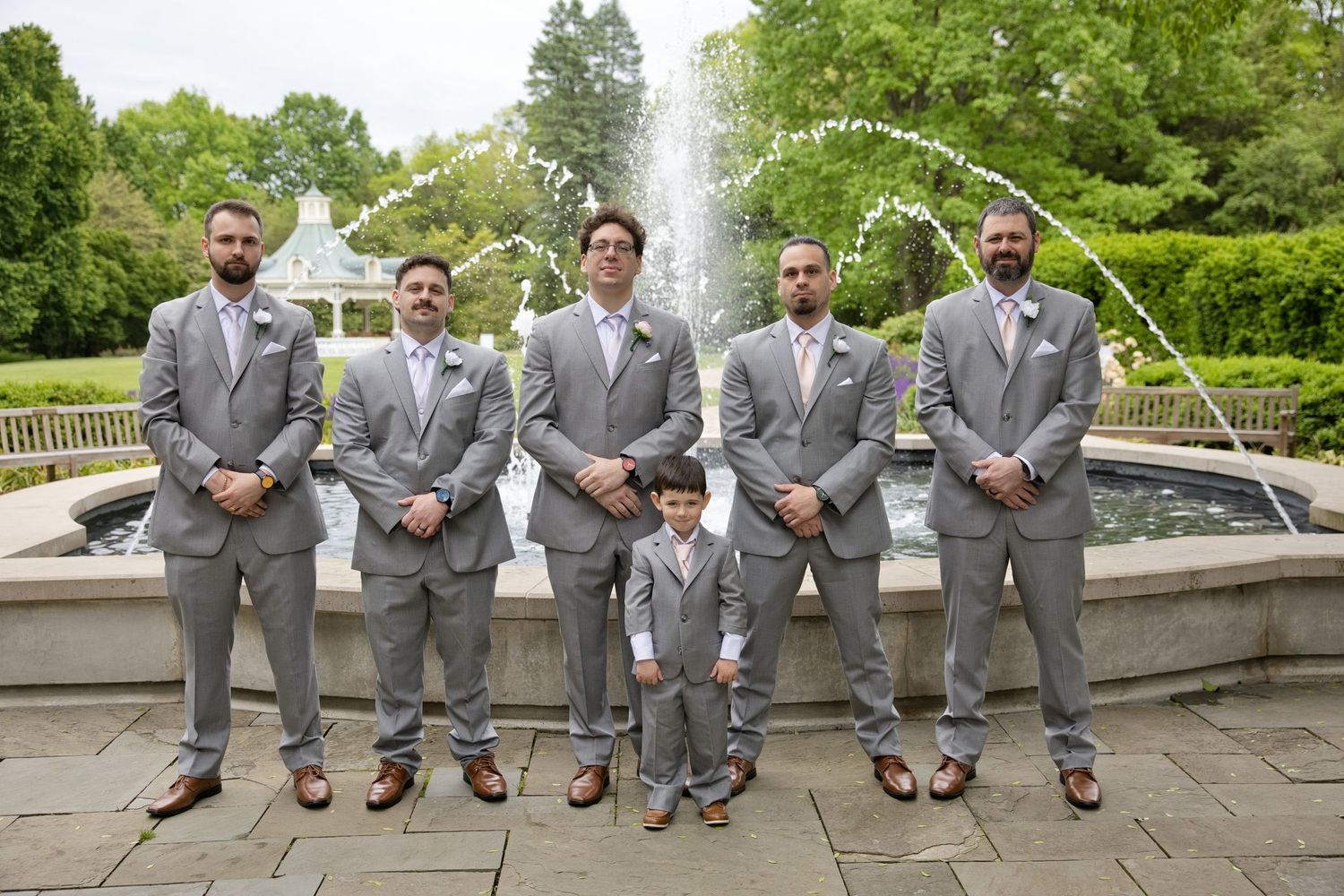 Groomsmen and ring bearer in gray suits pose in front of a garden fountain at an outdoor wedding ceremony.
