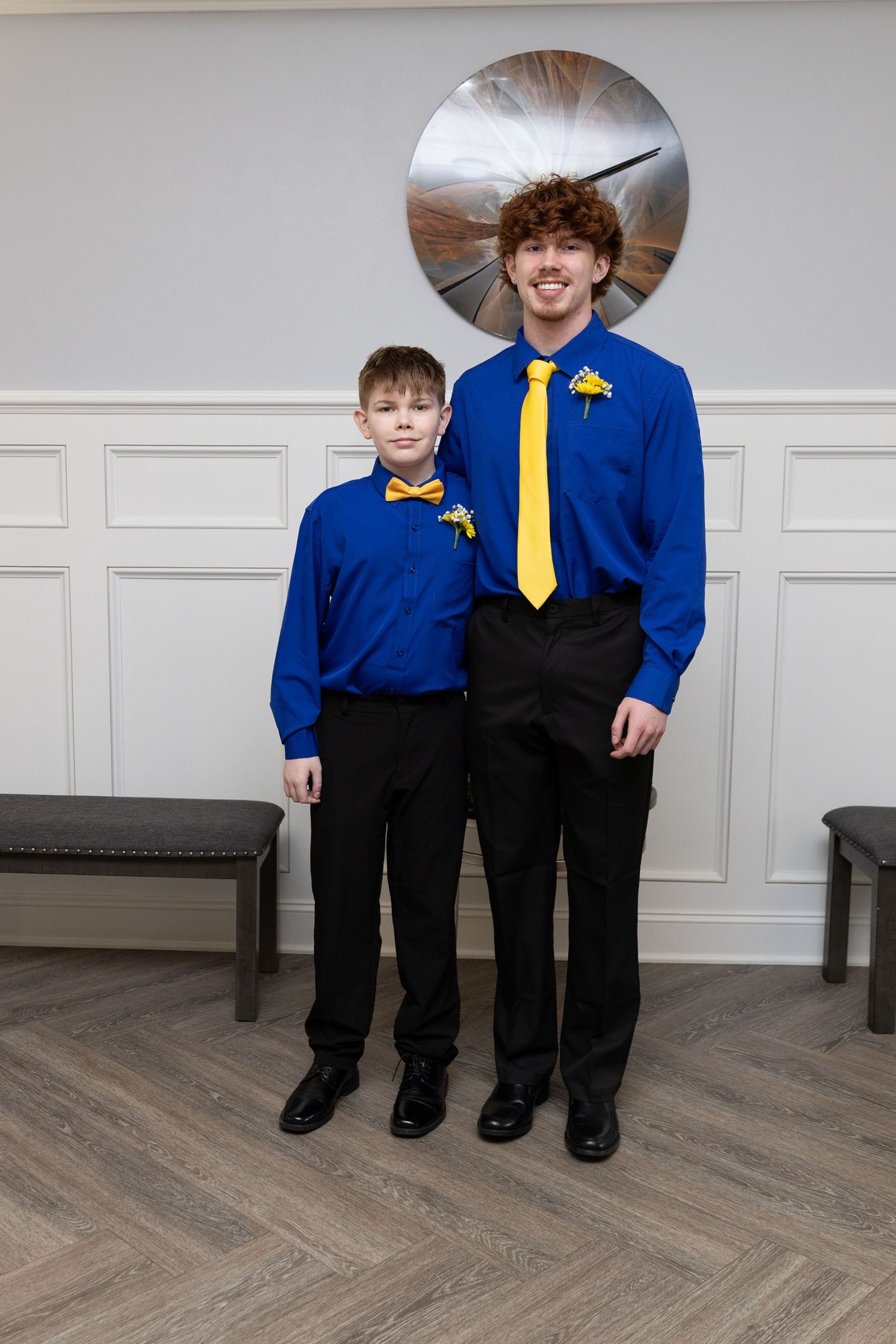 Two boys in matching royal blue shirts and black pants, wearing yellow bow tie and tie, posing indoors.
