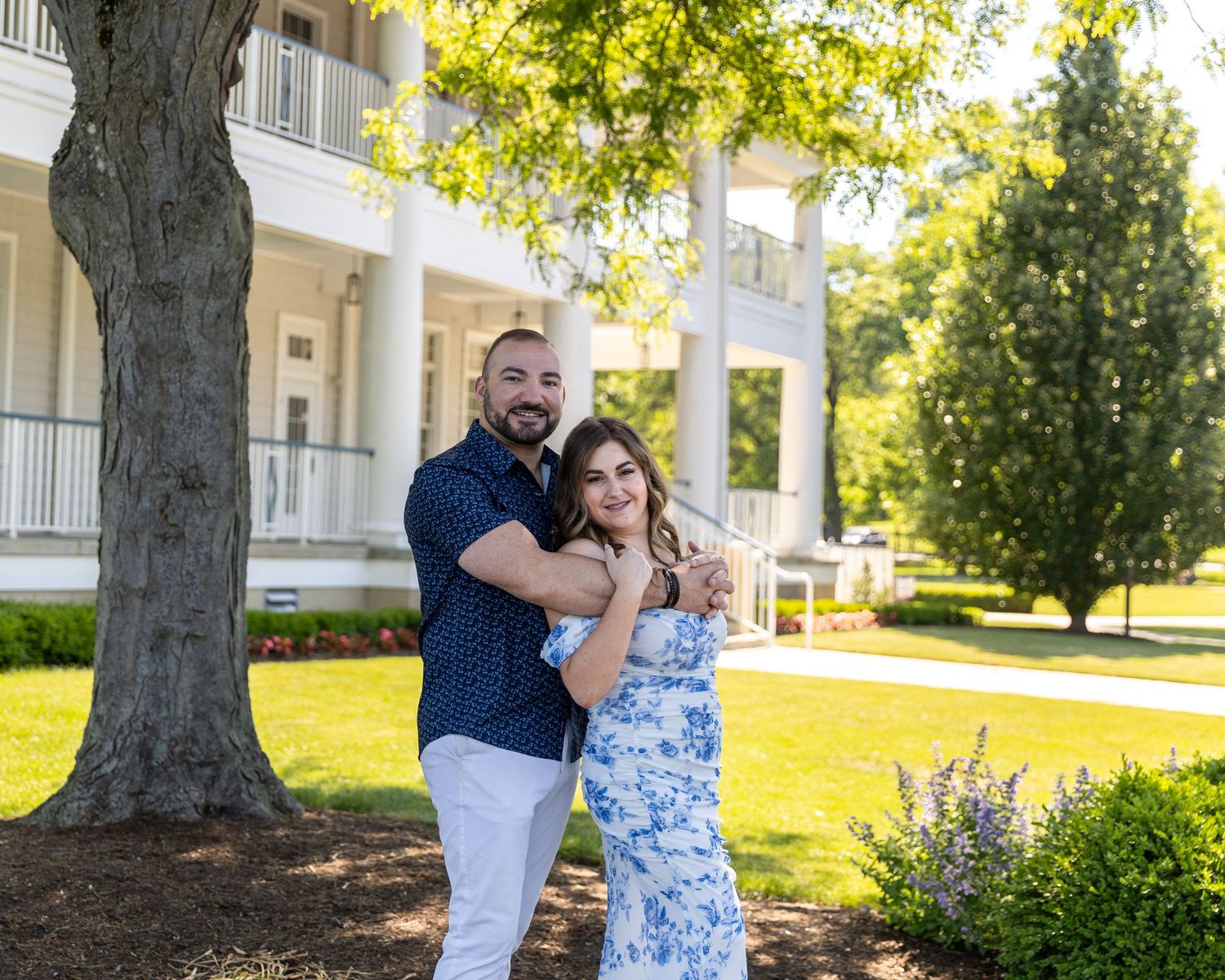 Couple embracing and smiling outdoors near a large tree with a white colonial mansion in the background on a sunny day.