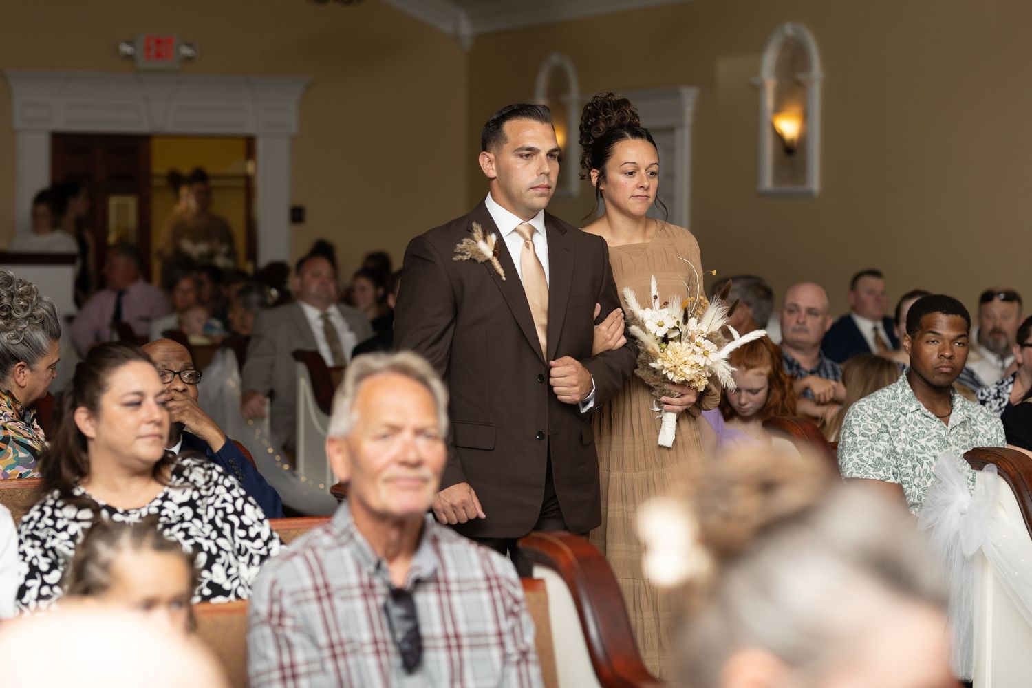 Wedding attendees fill the rows of seats in a formal indoor venue with warm lighting.