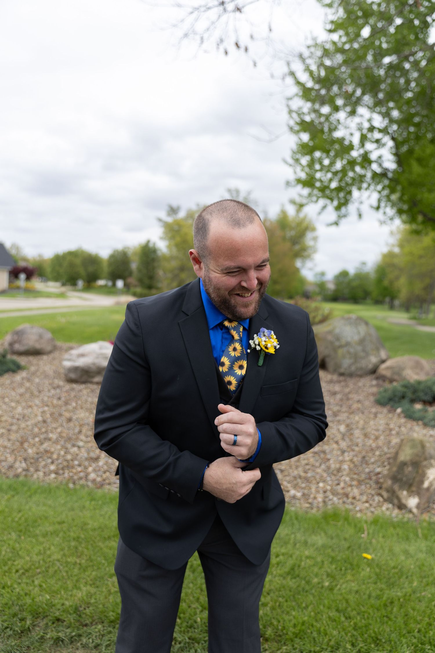 Smiling groom in black suit with blue shirt, sunflower tie and boutonniere, standing outdoors at a wedding venue.