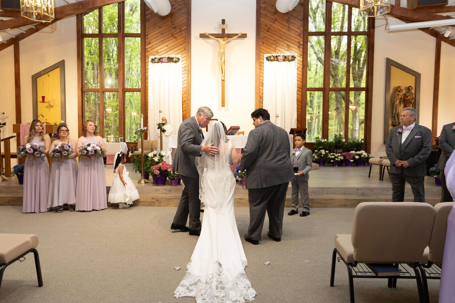 Bride and groom exchange vows at a church altar with bridesmaids in lavender dresses and a crucifix backdrop.