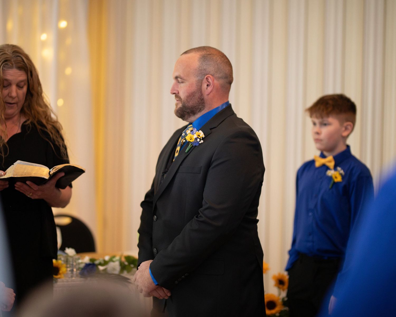 Groom in black suit with blue floral boutonniere stands during wedding ceremony while officiant reads from book.