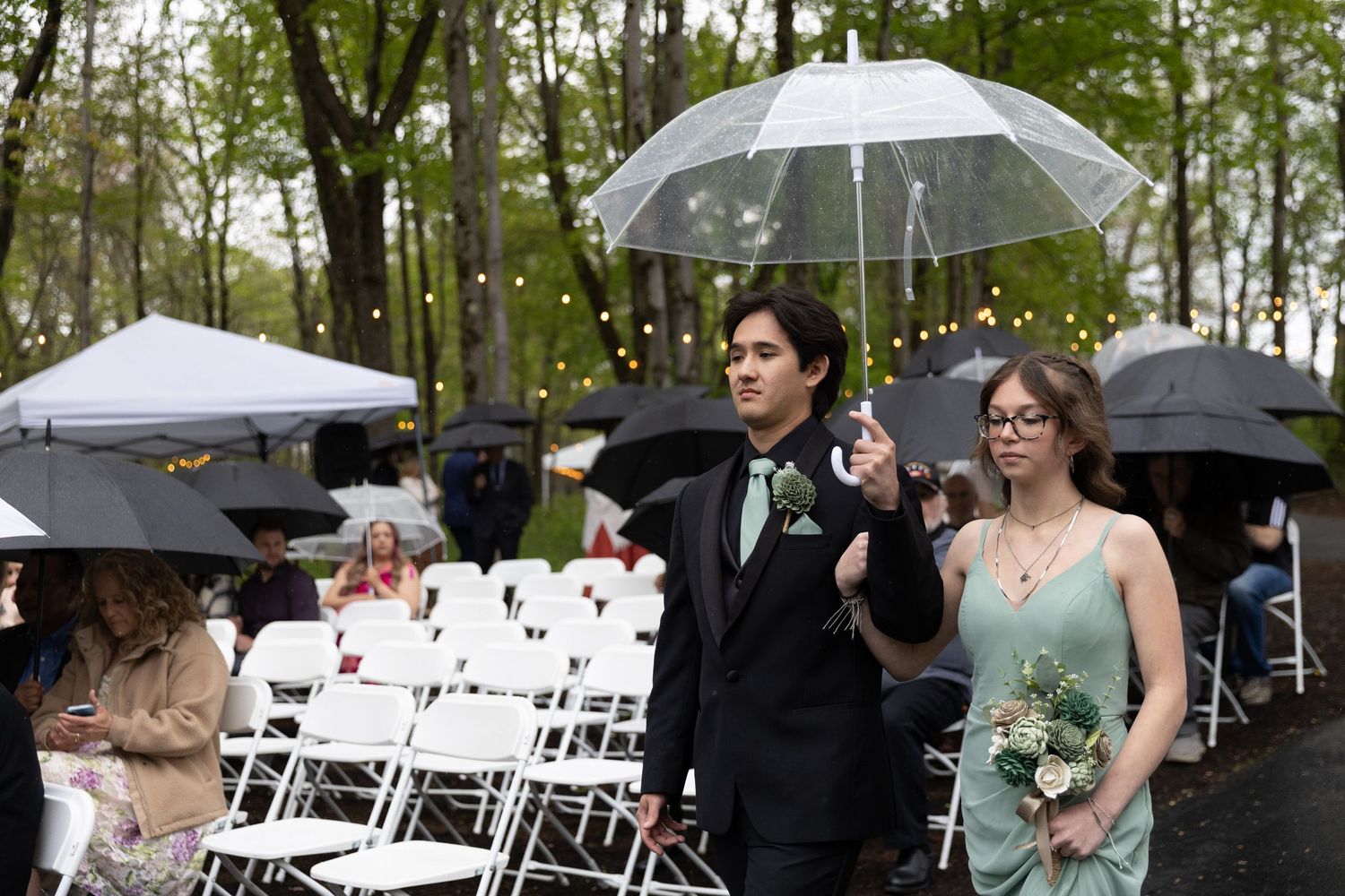 Groomsman escorts bridesmaid down outdoor wedding aisle in rain, holding clear umbrella among white chairs.
