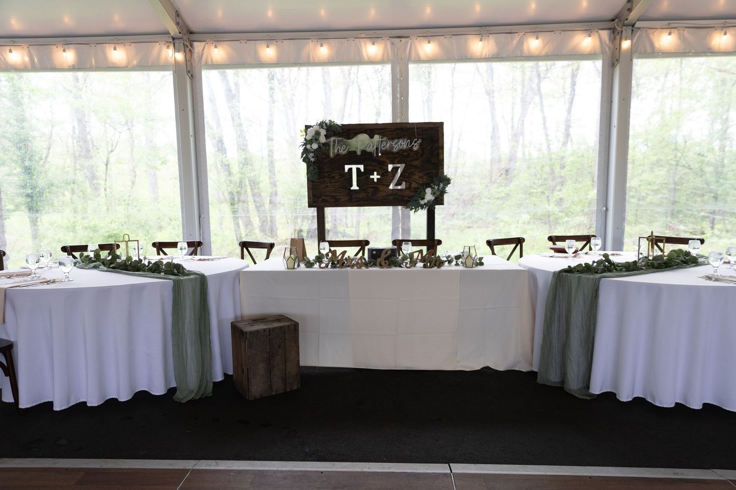 Elegant wedding head table with white linens, sage green runners, and a rustic wooden sign displaying T+Z initials under a tent.