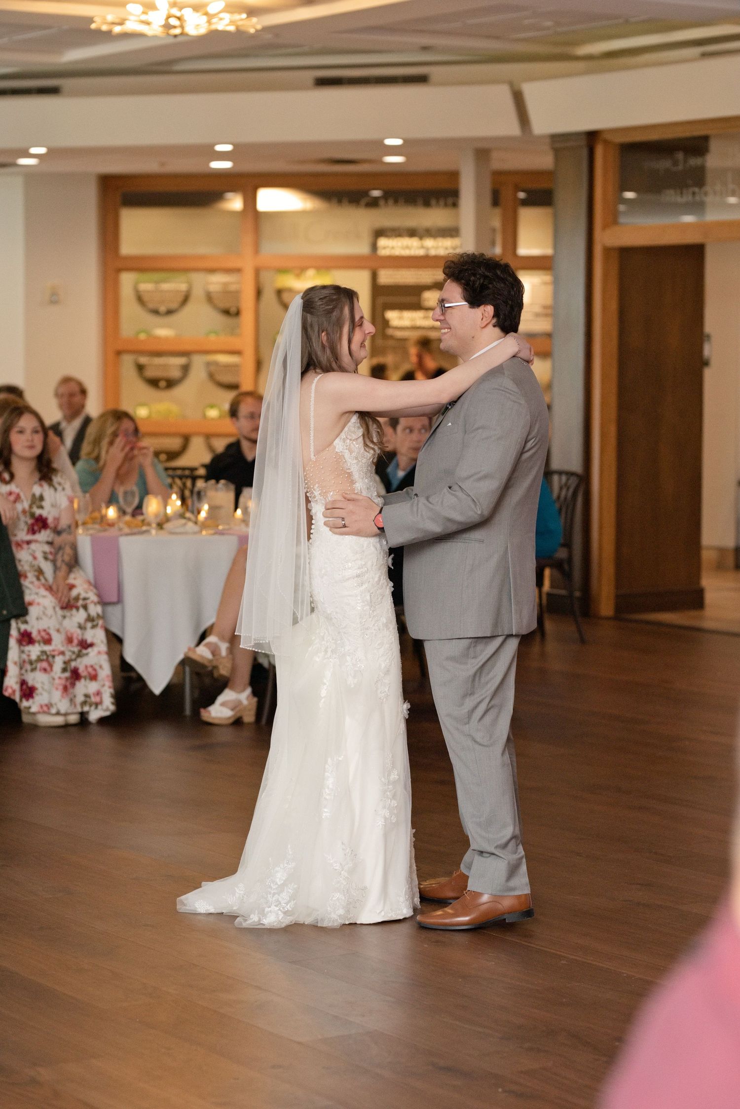 Bride and groom share their first dance at wedding reception, bride in lace gown with veil, groom in gray suit.
