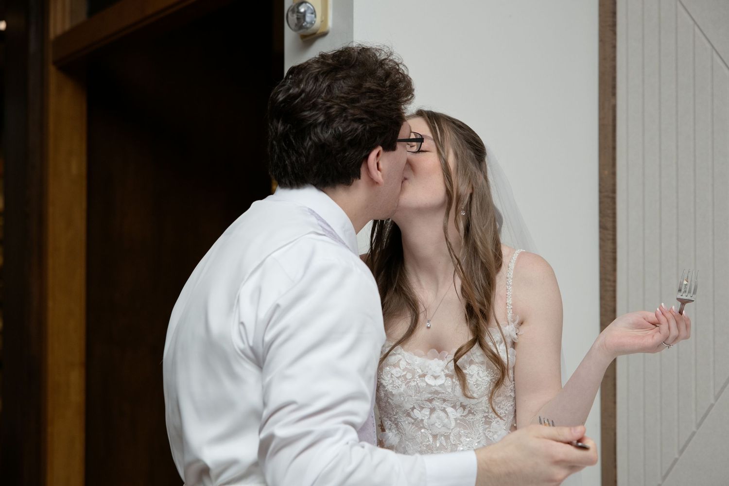 Newlyweds sharing a romantic kiss after cutting their wedding cake, bride holding a fork.