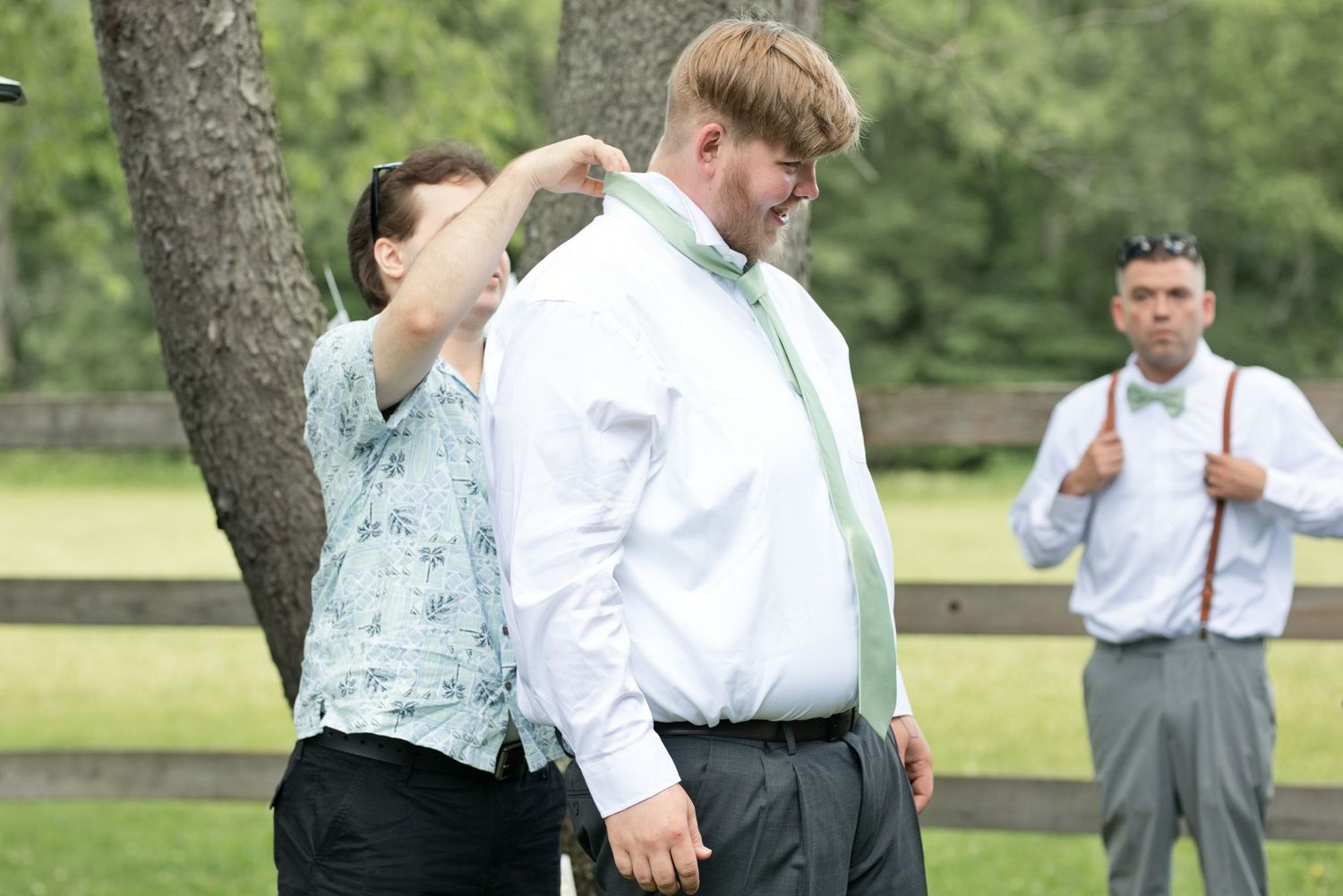 Woman helping groom with his green tie outdoors under a tree before a wedding ceremony.