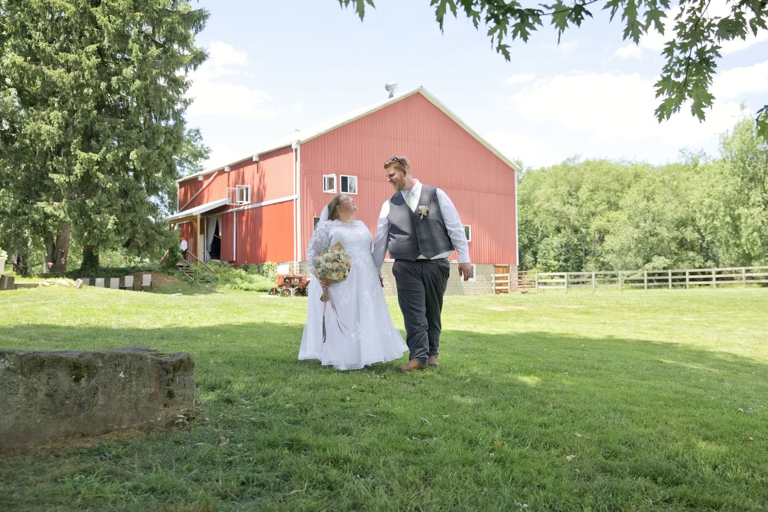 Bride and groom walking hand in hand on green lawn beside a red barn on their wedding day.