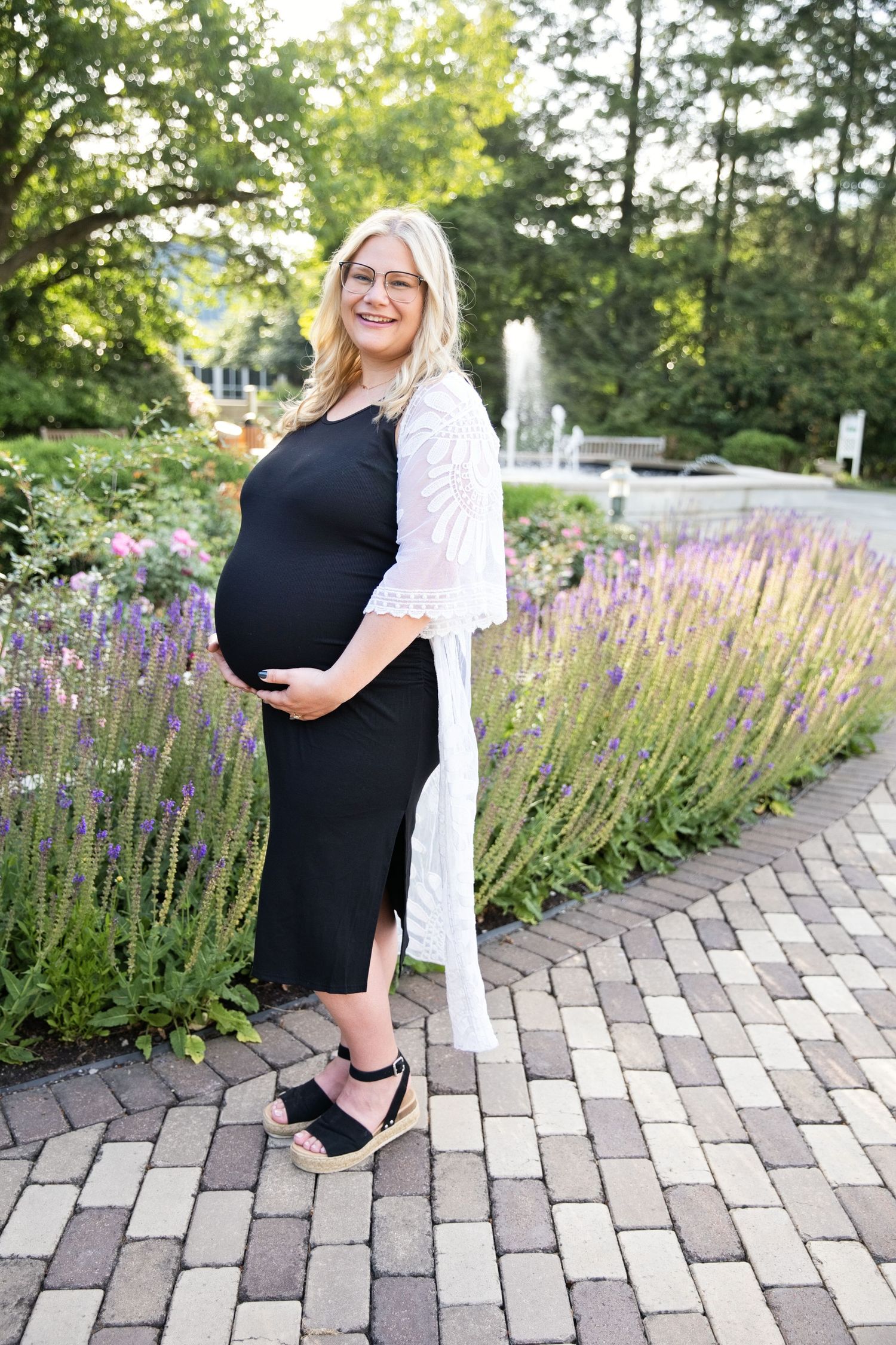 Pregnant woman in black dress and white lace cardigan stands among purple lavender flowers on brick path.