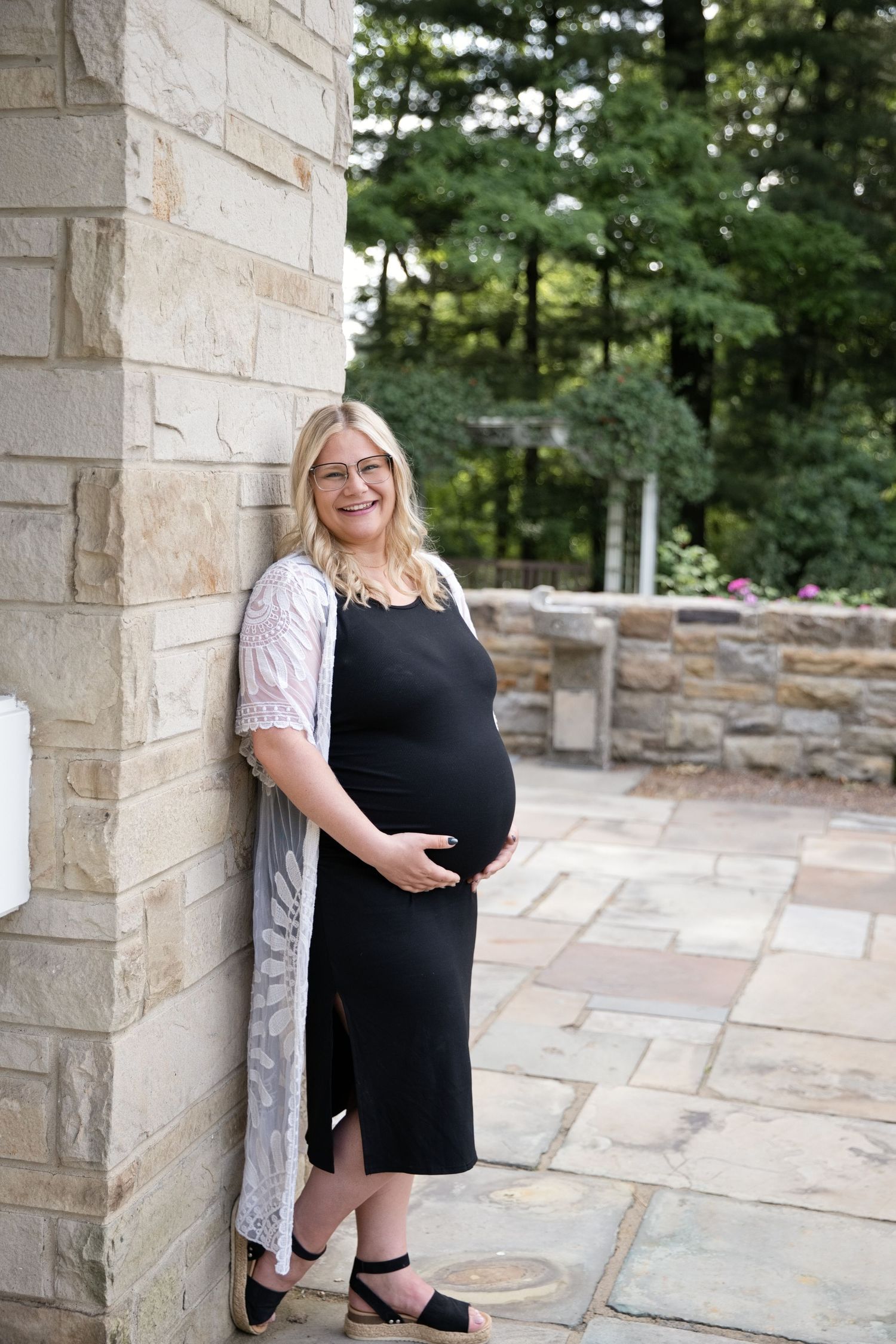 Smiling pregnant blonde woman in black dress and white lace cardigan leans against stone wall outdoors.