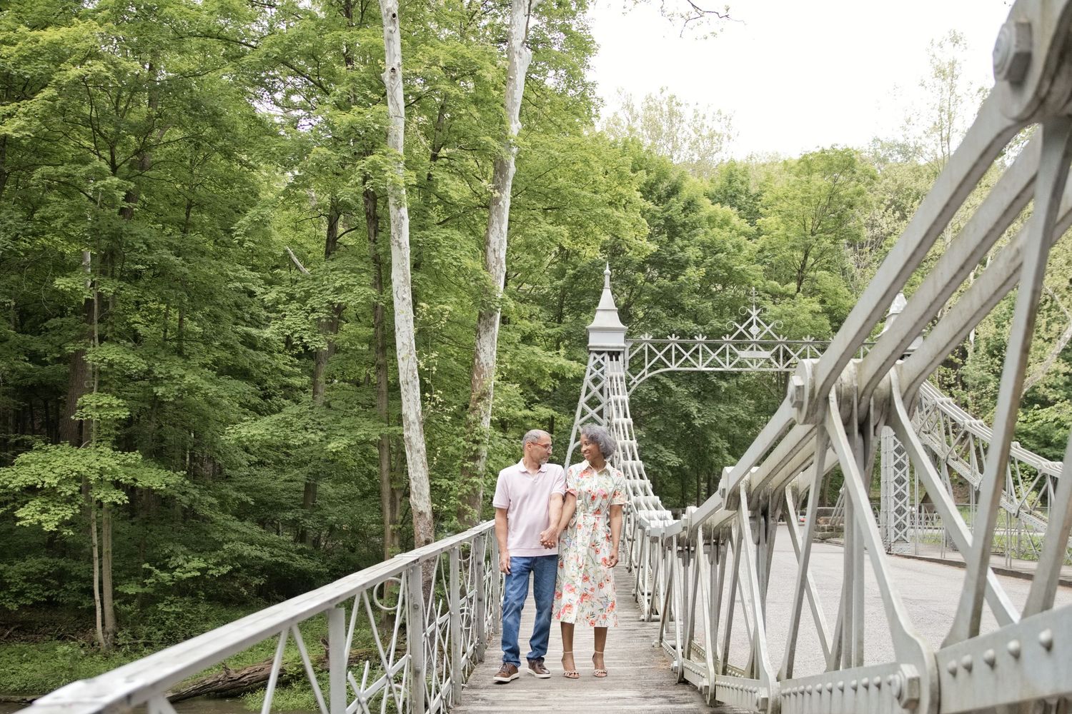 Couple walking hand-in-hand on a white metal suspension bridge surrounded by lush green trees.