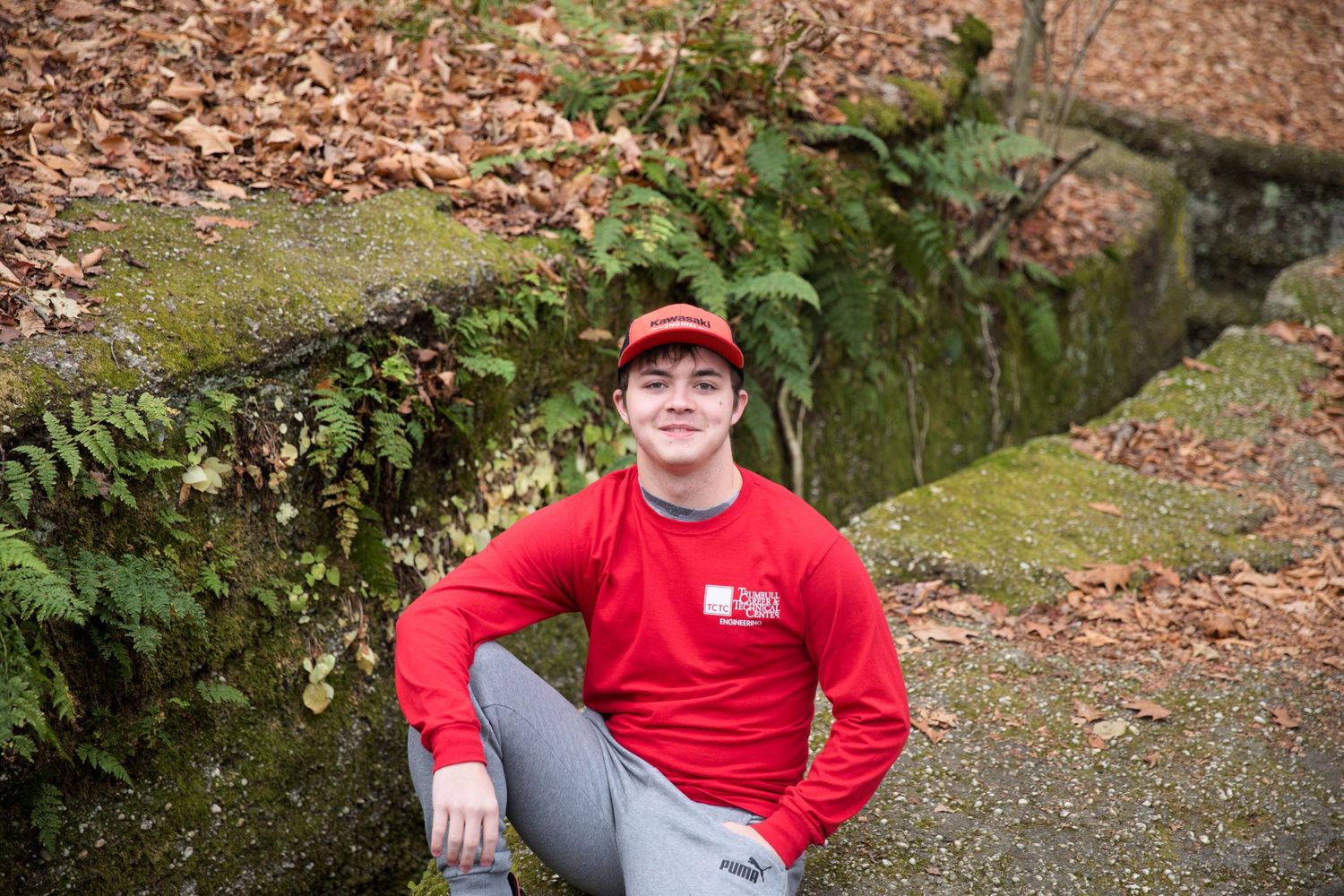 People in red shirts exploring a wooded hiking trail with fallen leaves and mossy rocks.