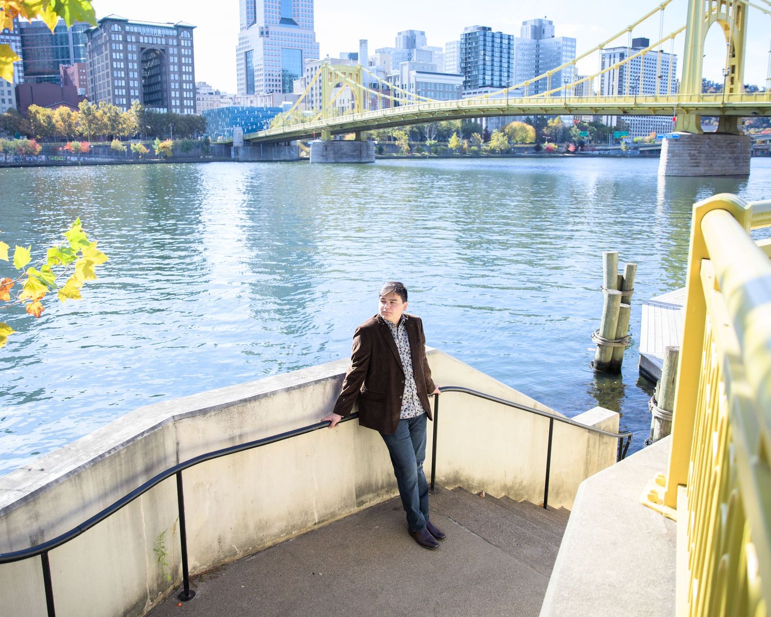 Yellow bridge spans across river with downtown Pittsburgh skyline and skyscrapers visible in background.