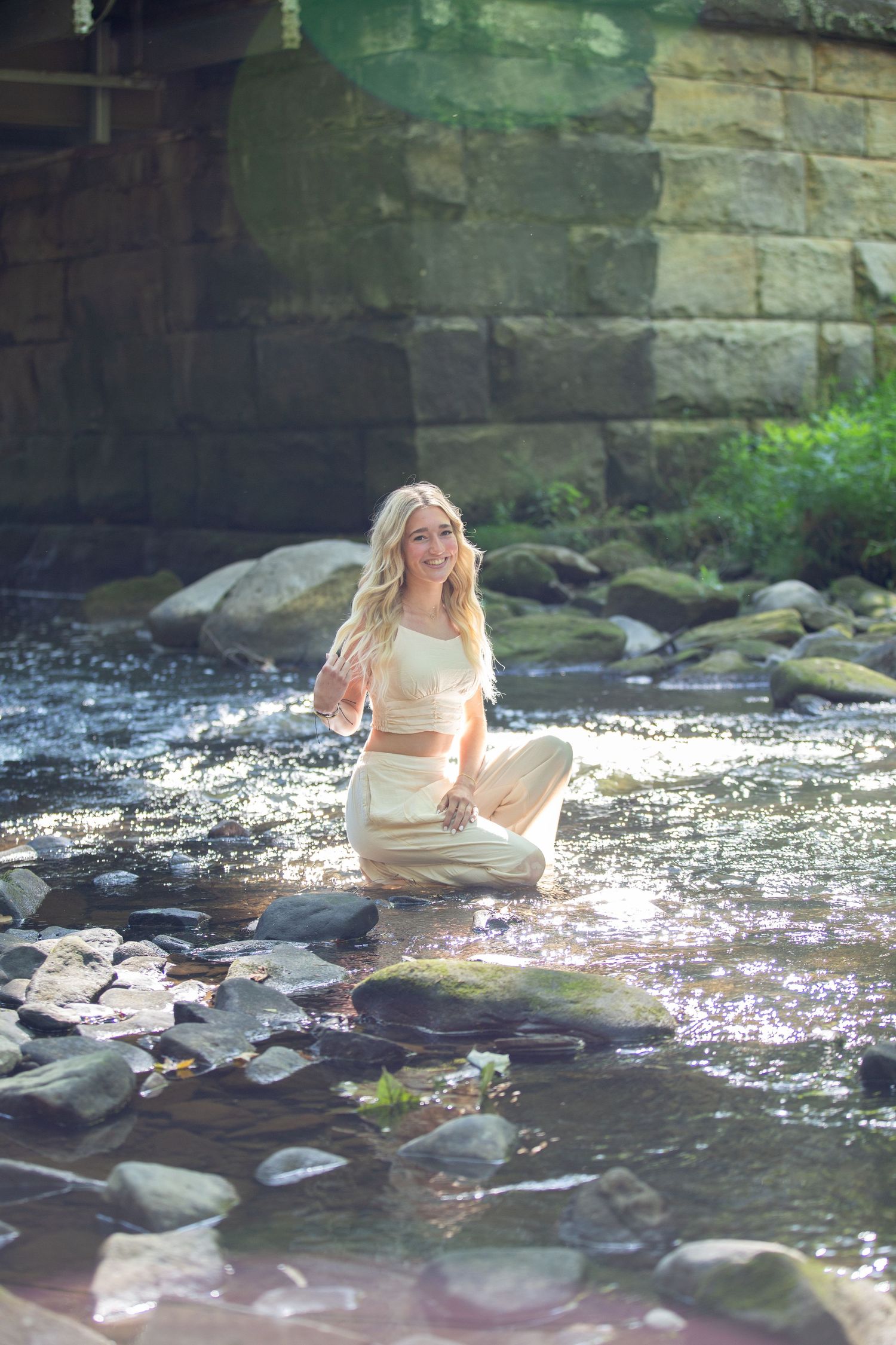 Person in white dress sits on rocks by flowing stream with sunlight sparkling on the water.