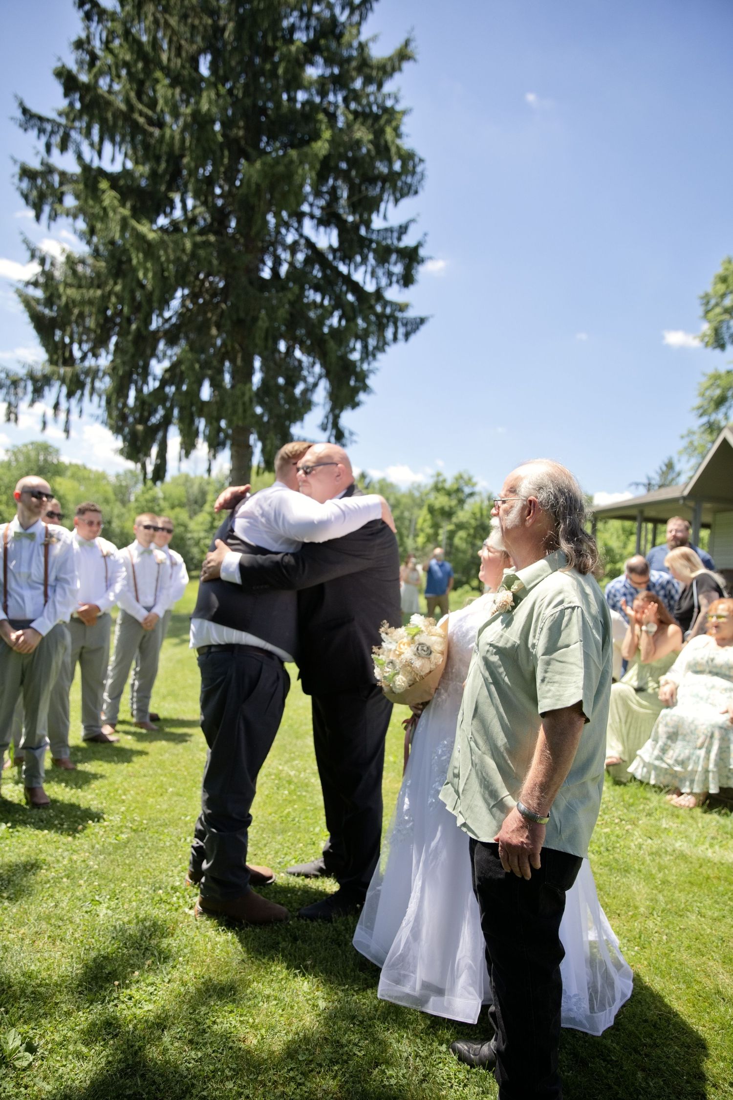 Outdoor wedding ceremony on green lawn with groom embracing guest, bride holding bouquet, guests seated, tall evergreen tree backdrop.