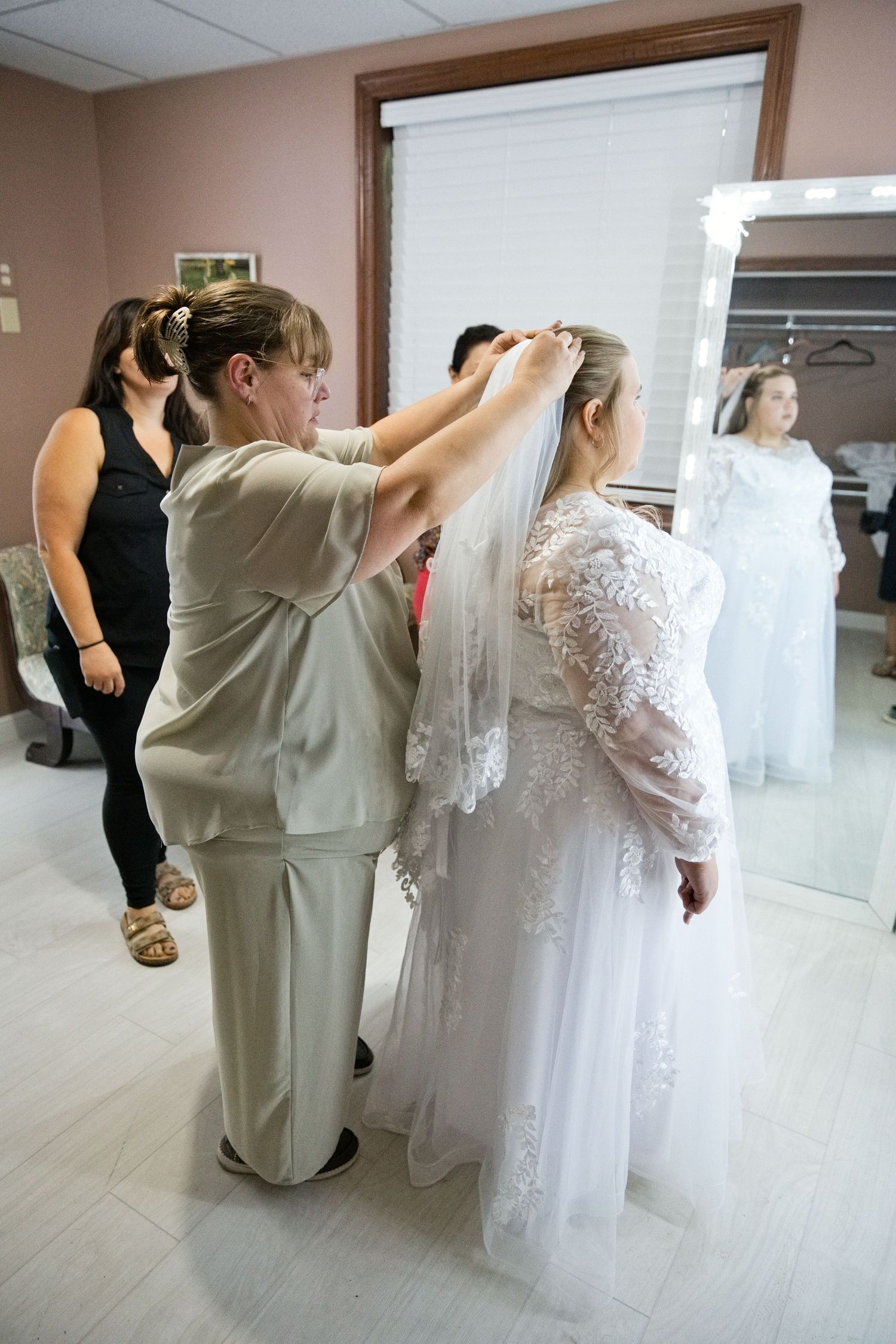 A bride in a white lace gown has her veil adjusted by a woman in a bridal boutique fitting room.