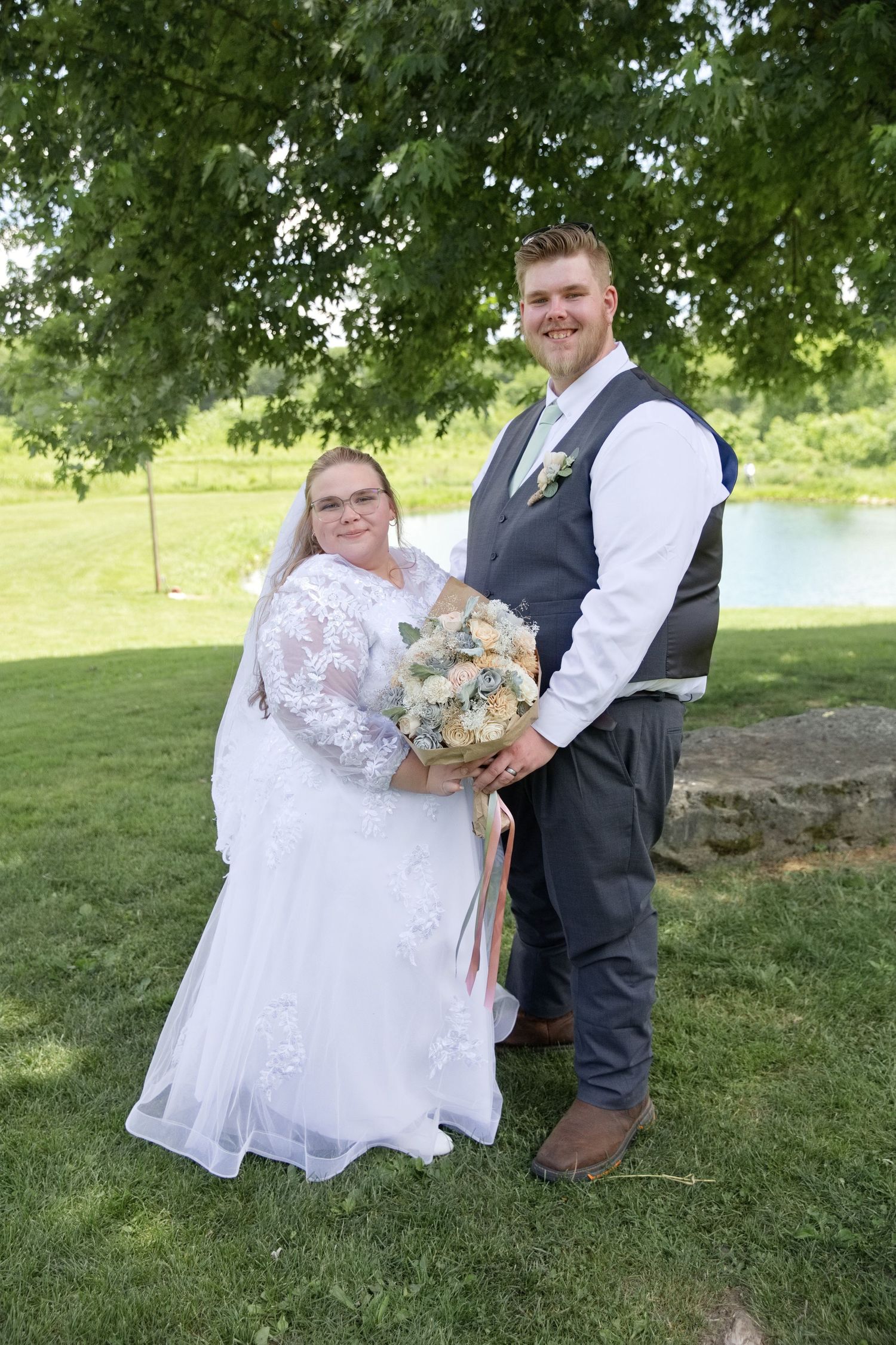 Bride in lace gown holding bouquet stands with groom in gray vest outdoors under green trees near pond.