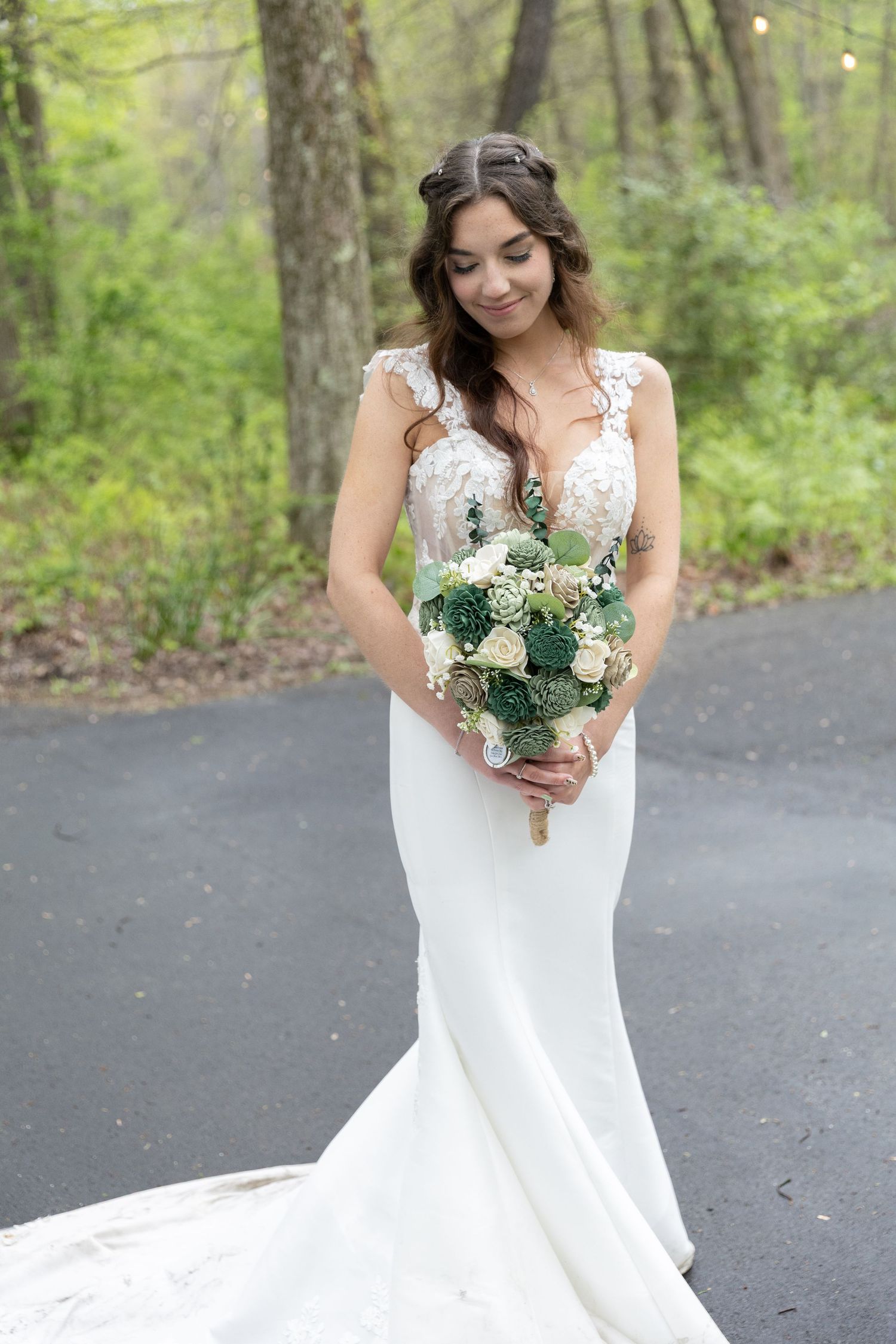 Bride in white lace mermaid gown holding green and white bouquet, standing on a tree-lined path.