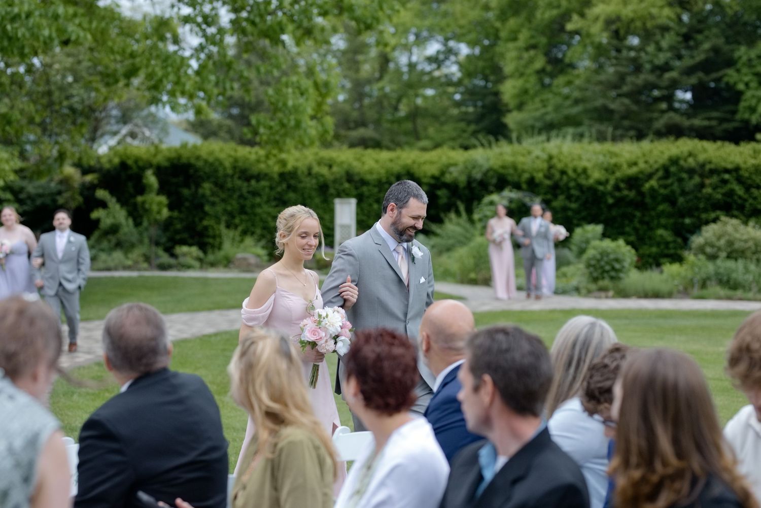 Bride and groom walk down outdoor wedding aisle surrounded by guests in a lush garden setting.