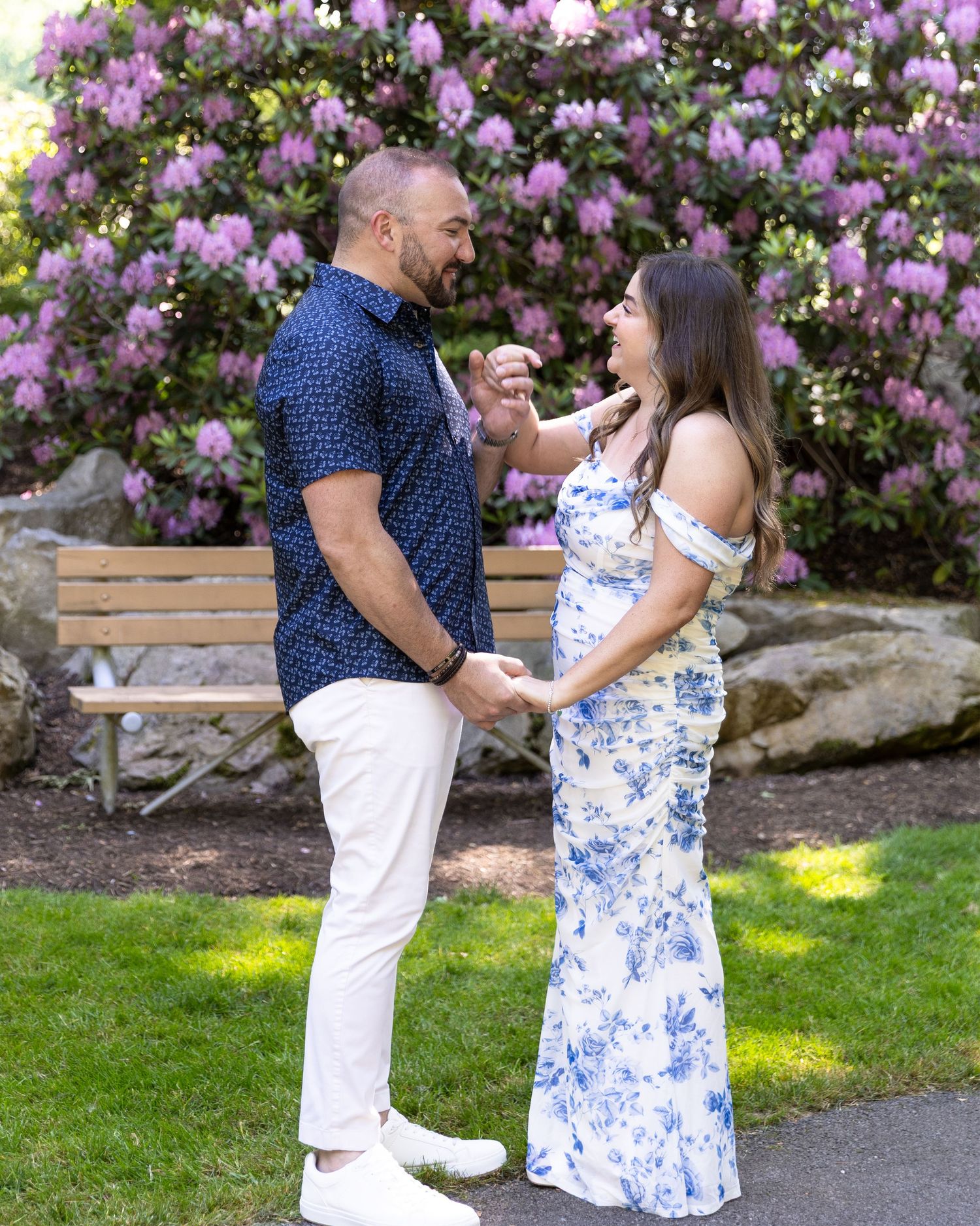 Couple laughing and holding hands in front of purple rhododendrons on a sunny day in a lush garden.