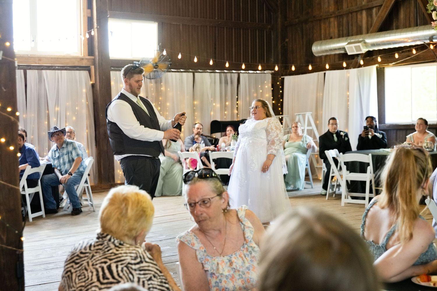 Rustic barn wedding reception with couple standing near guests seated in white chairs under string lights.