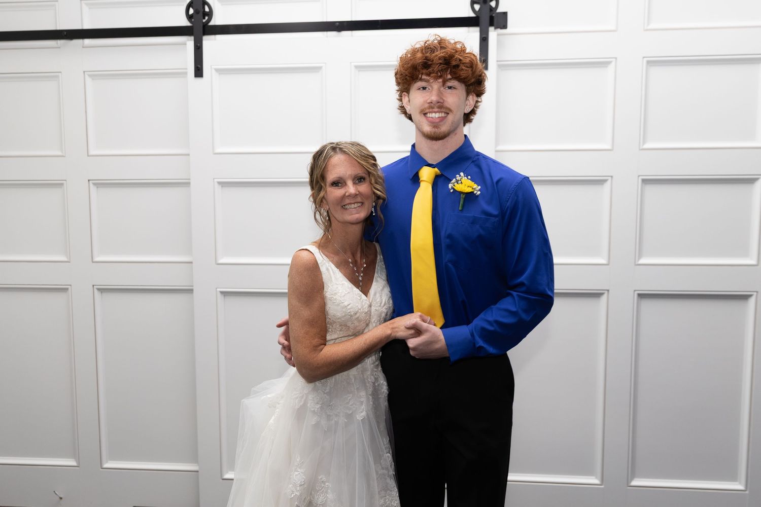 A woman in a white wedding dress poses with a young man in a blue suit and yellow tie in front of white barn doors.