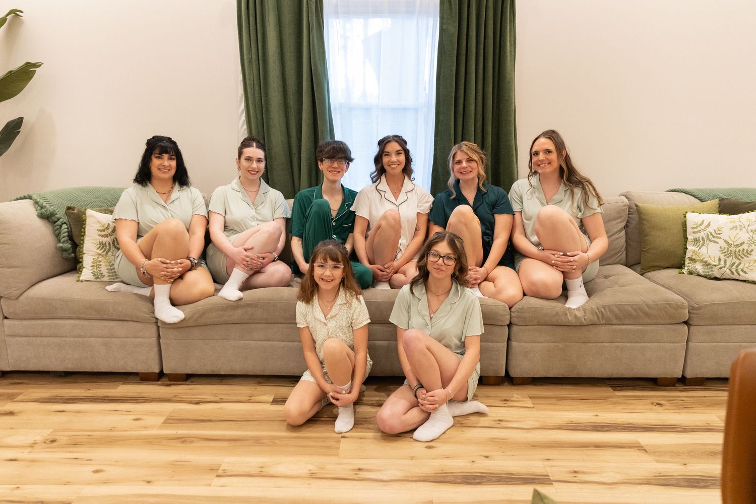Group of nine women in matching green and white pajamas posing together on a beige sectional sofa indoors.