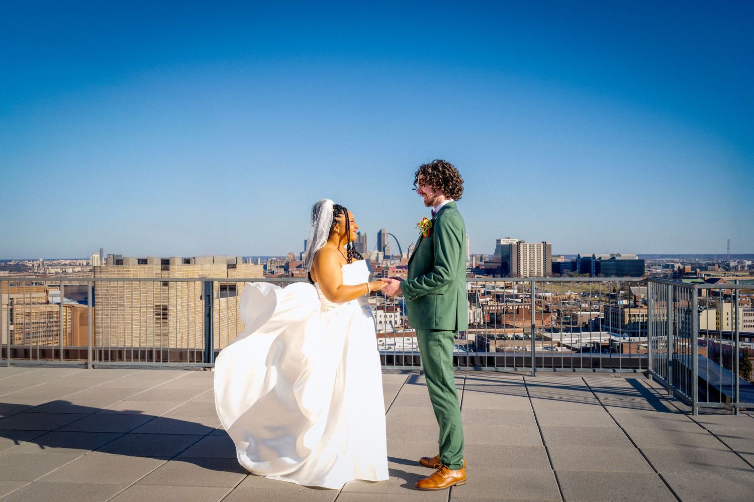 Bride and groom facing each other on urban rooftop, bride in flowing white gown, groom in green suit, blue sky backdrop.
