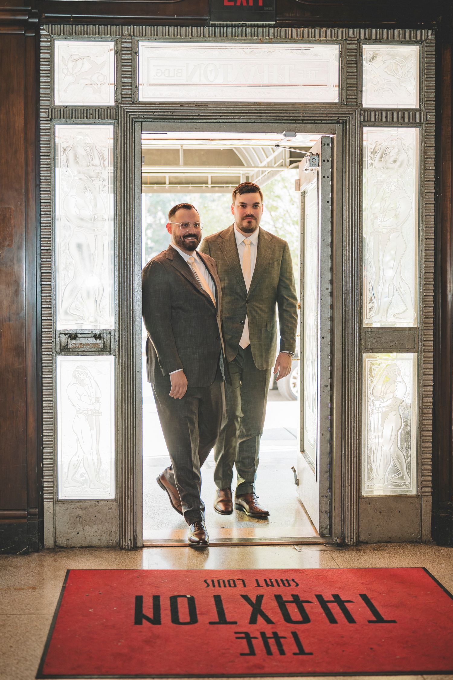 Two grooms in matching gray suits enter The Thaxton Saint Louis venue through ornate glass doors.