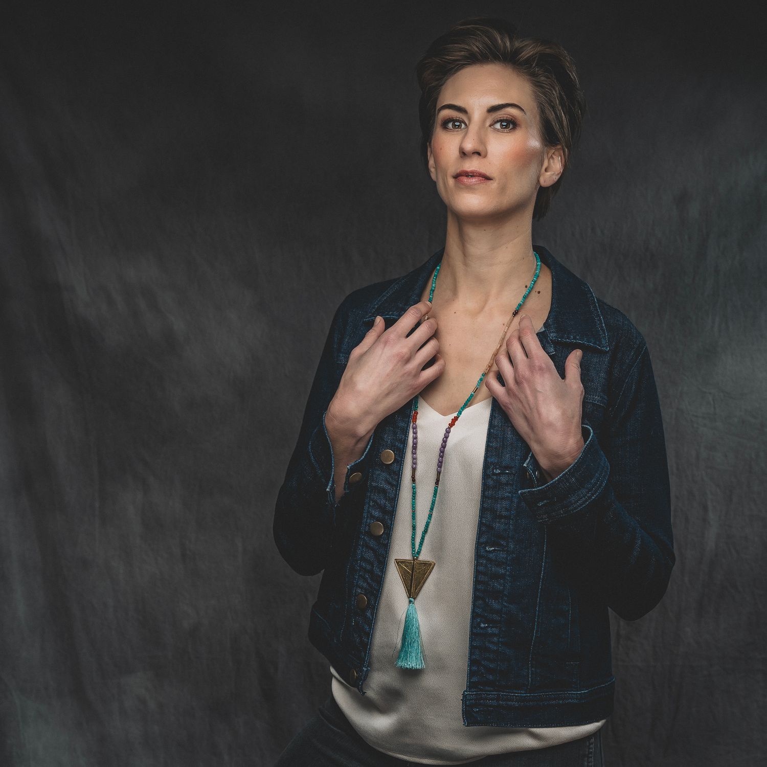 Woman in a denim jacket holds a tassel necklace, posing against a dark textured studio backdrop.