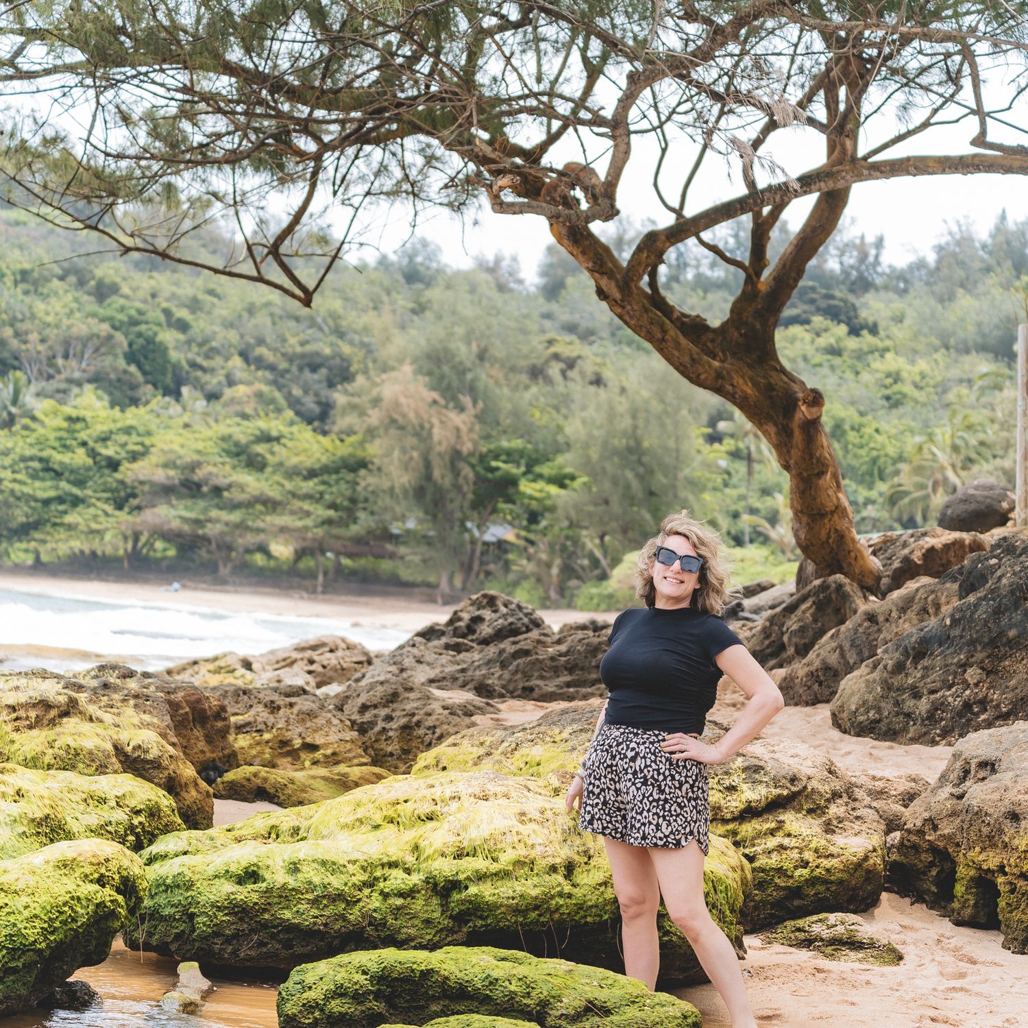 Woman in black top and leopard print skirt poses on mossy rocks at a tropical beach with lush green trees.