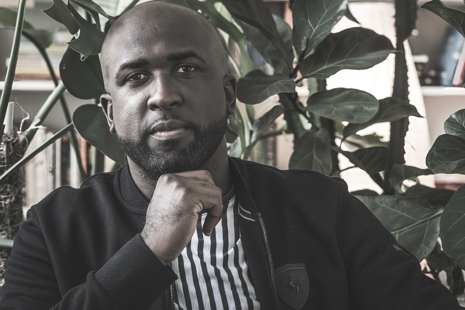 Bald man with a beard sits thoughtfully among tropical green plants in a moody, candid portrait.