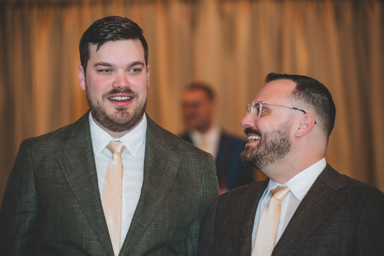 Two groomsmen in gray suits share a joyful moment smiling at each other at a wedding reception.