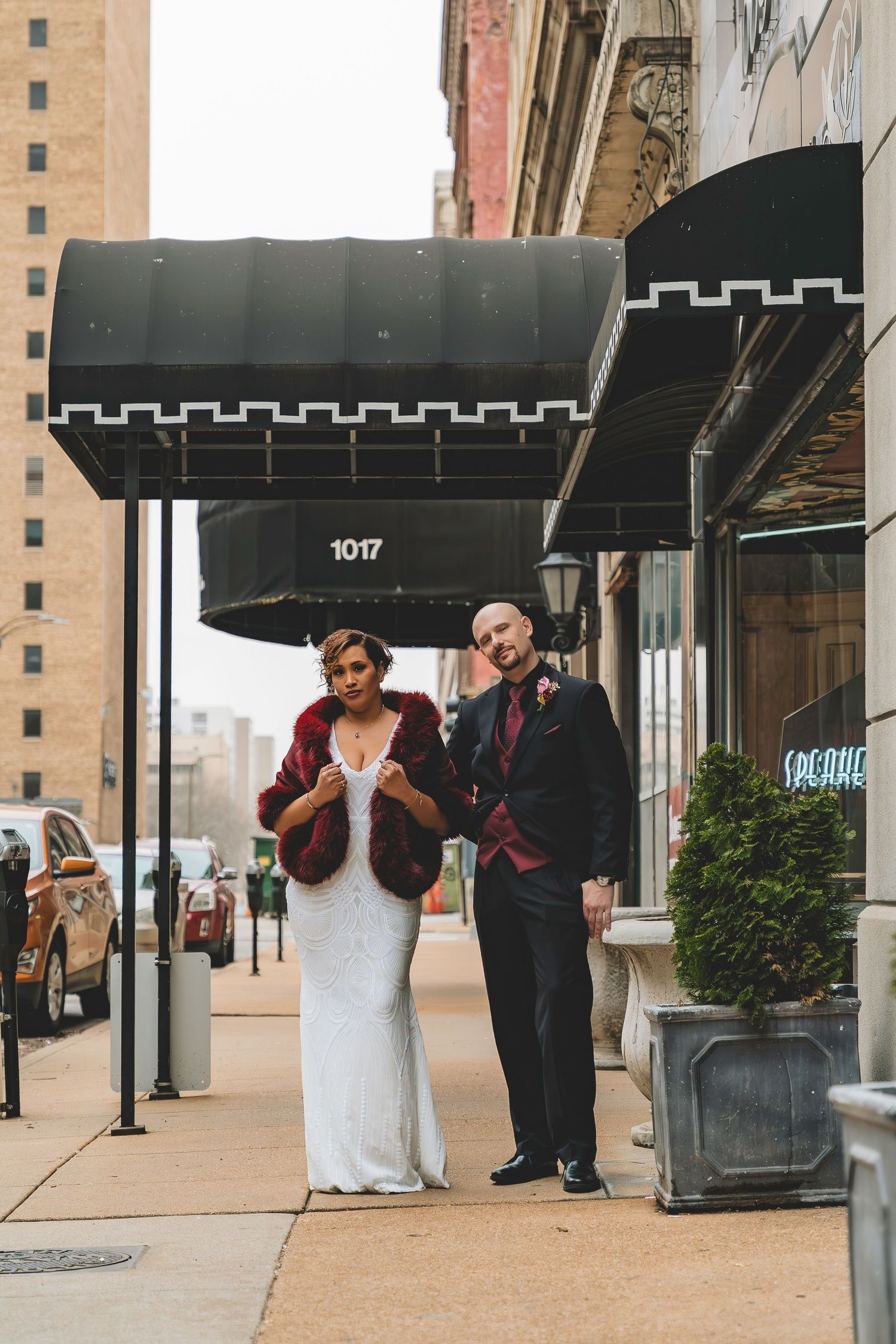 Bride in white beaded gown with fur stole and groom in dark suit pose on urban sidewalk under black awning.