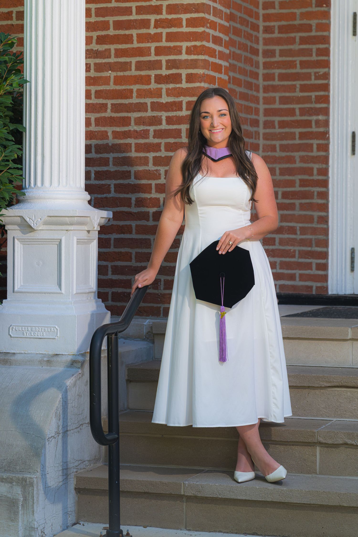 Brunette woman in white dress holds graduation cap on brick building steps.