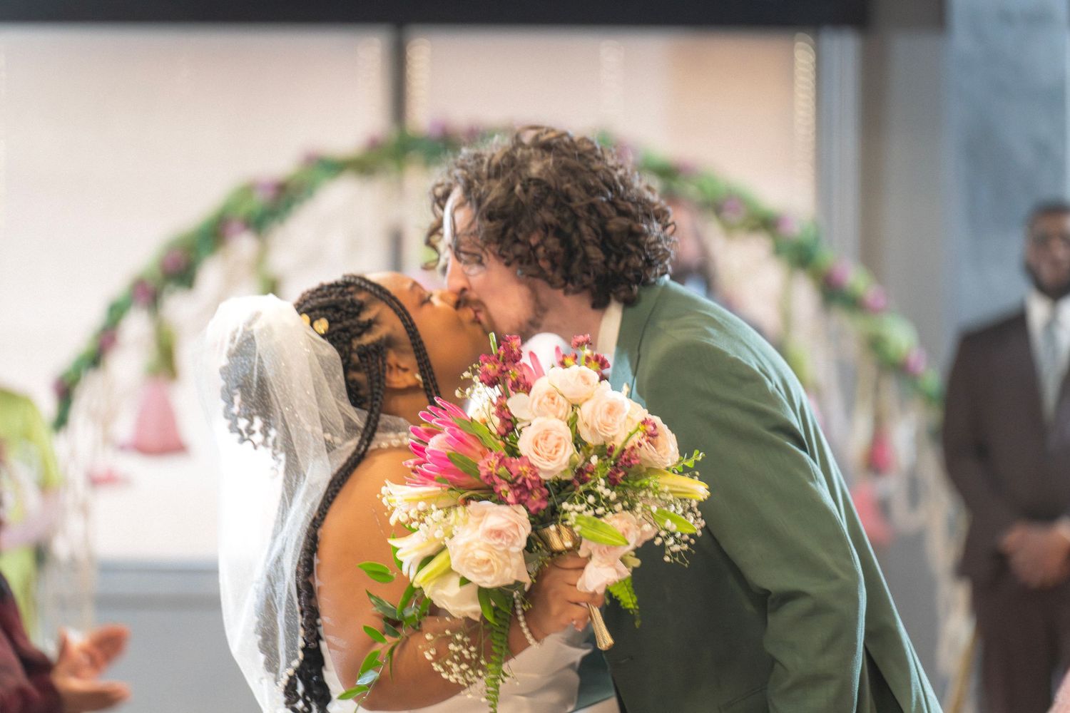 Newlyweds sharing their first kiss at the altar, bride holding colorful bouquet, decorative floral arch in background.